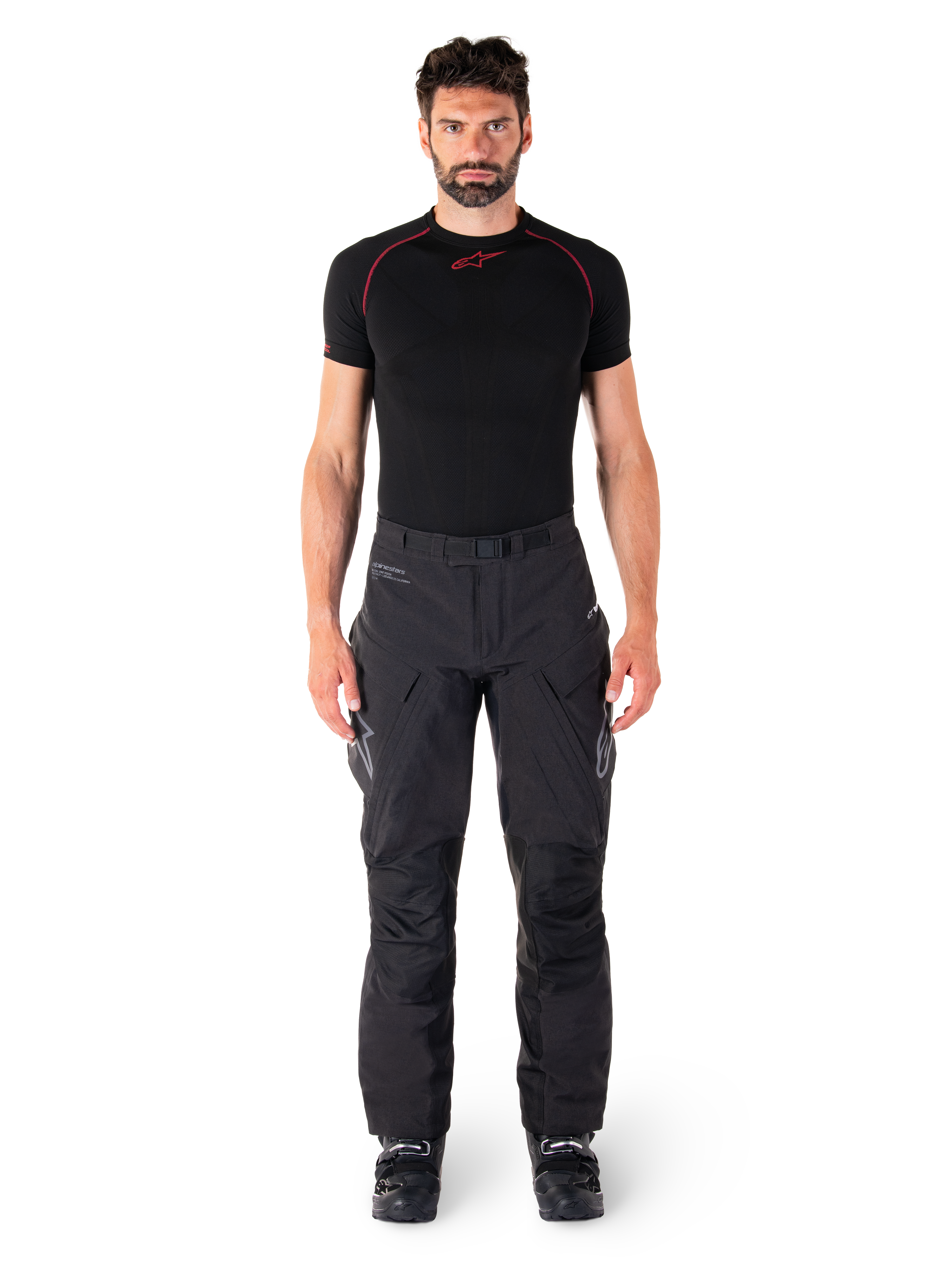 Calças Alpinestars Hyde XT Drystar®, calças de motociclismo Adventure Touring, pretas, vestidas por um modelo com uma camisola interior de manga curta preta e botas pretas, com material elástico técnico, painéis reforçados nos joelhos e bolsos de carga ergonómicos.