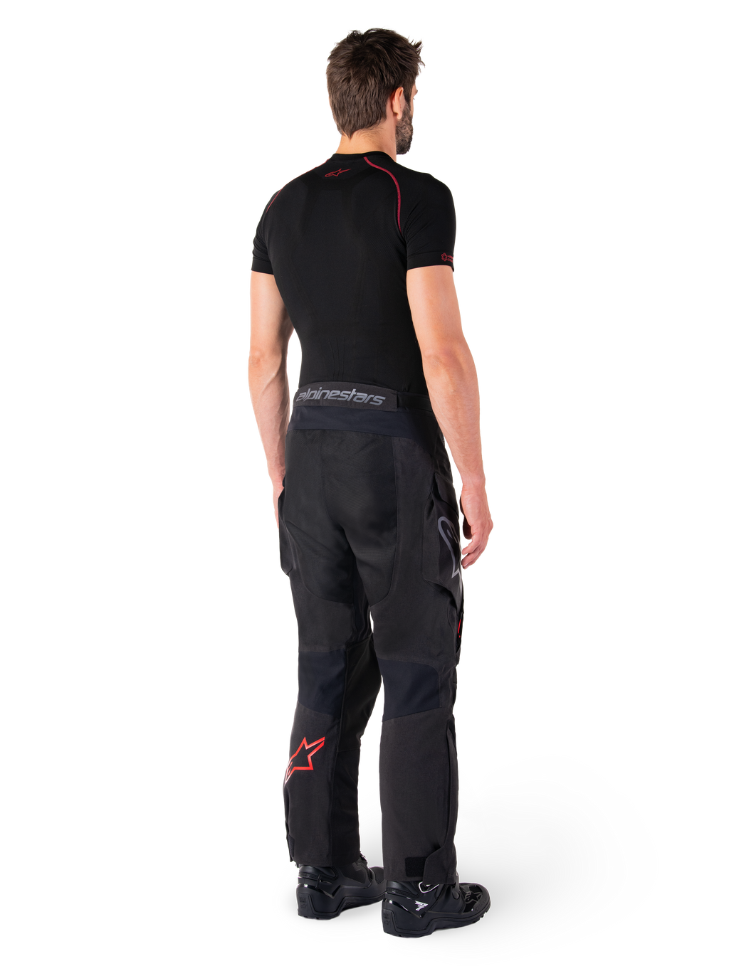 Calças Alpinestars Hyde XT Drystar®, calças Adventure Touring, pretas e vermelho brilhante, vista traseira de um piloto a usar as calças com uma camada base preta e botas pretas, apresentando construção têxtil durável com um logótipo de estrela vermelha na parte inferior da perna e marca Alpinestars cinzenta no cós.