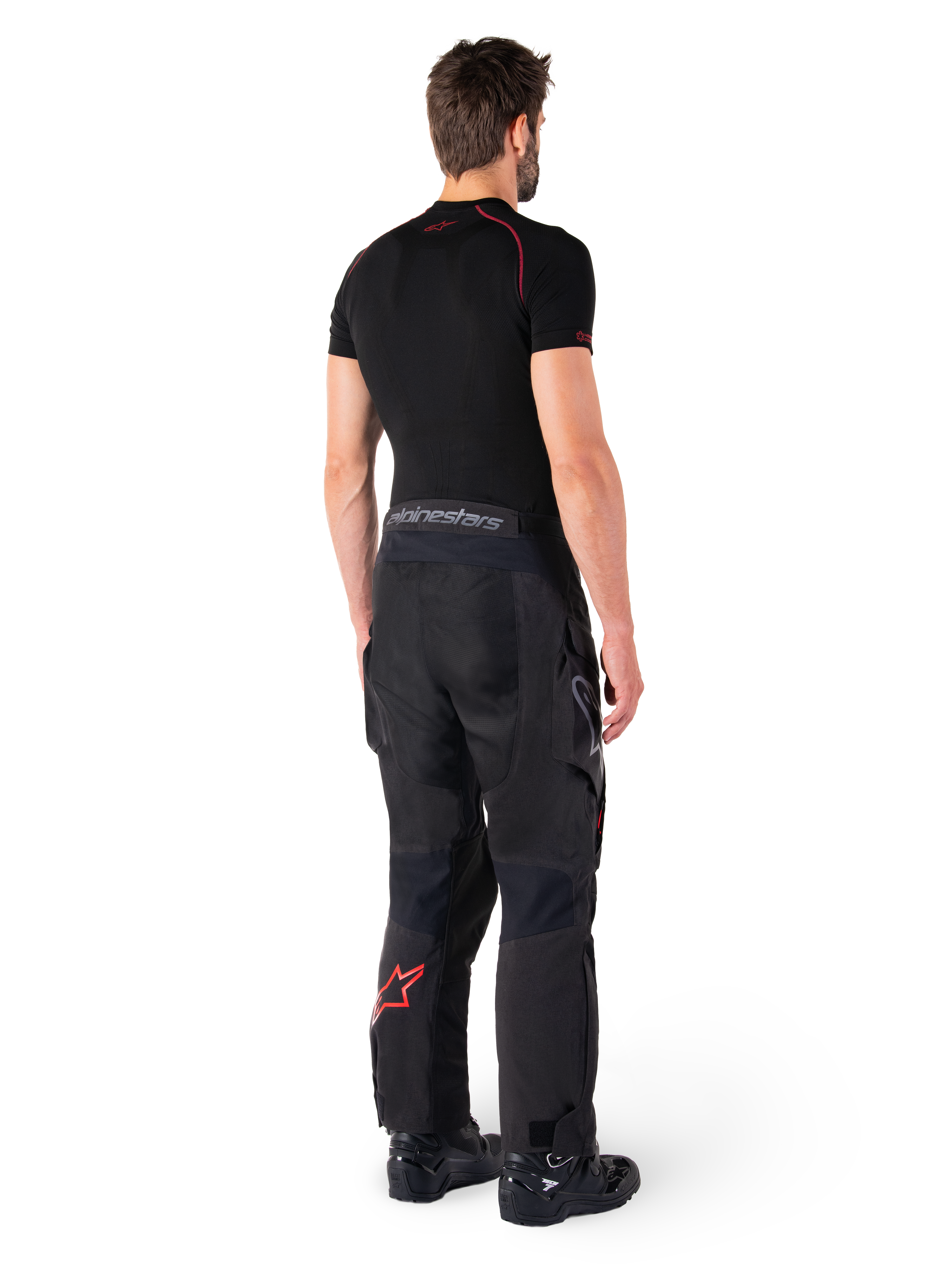 Calças Alpinestars Hyde XT Drystar®, calças Adventure Touring, pretas e vermelho brilhante, vista traseira de um piloto a usar as calças com uma camada base preta e botas pretas, apresentando construção têxtil durável com um logótipo de estrela vermelha na parte inferior da perna e marca Alpinestars cinzenta no cós.