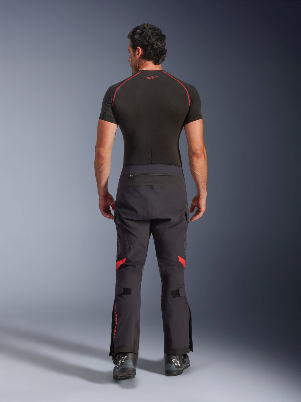 Calças Alpinestars Halo Pro Drystar® XF Laminated, calças de motociclismo de aventura, Preto Dark Shadow com detalhes em vermelho, vista traseira, usadas com uma camisola interior de manga curta preta e botas de aventura pretas, construção laminada de 3 camadas e ajuste ergonómico.
