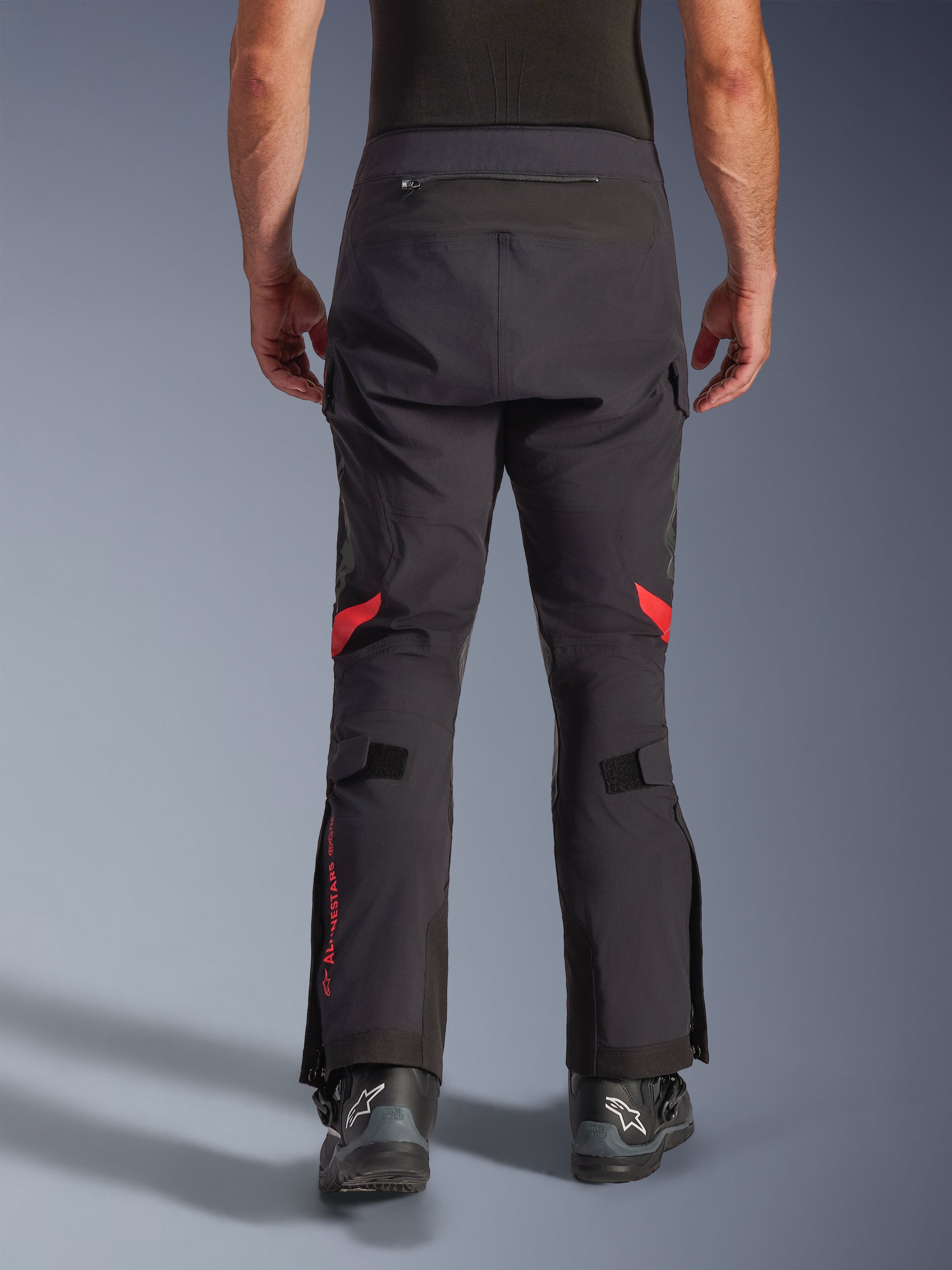 Alpinestars Halo Pro Drystar® XF Laminated Pants, Black Dark Shadow, vista traseira de calças de motociclismo de aventura com detalhes vermelhos atrás dos joelhos e logótipo vermelho na panturrilha, usadas com botas pretas Alpinestars, têxtil laminado 3L durável para desempenho todo-o-terreno