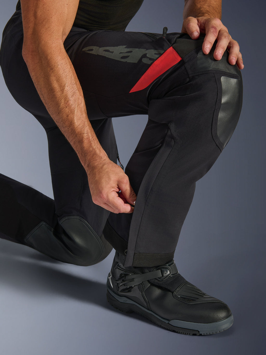 Calças Alpinestars Halo Pro Drystar® XF Laminated em Preto Dark Shadow, primeiro plano da parte inferior da perna com fecho de correr ajustável na panturrilha, reforços duráveis nos joelhos, detalhes em vermelho e marca 'astars', usadas com botas de motociclismo pretas.