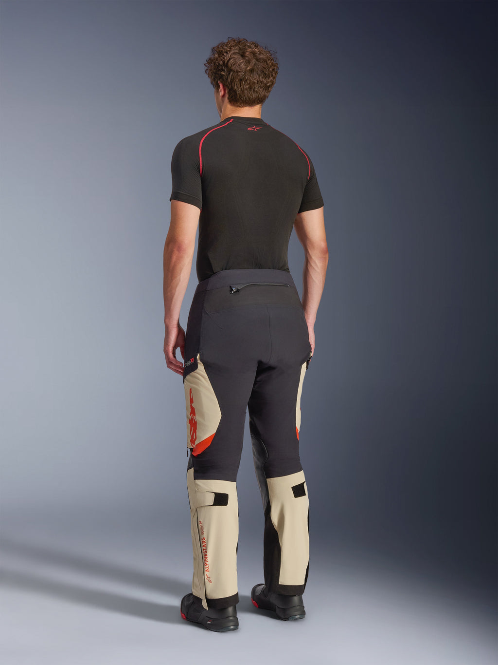 Calças Alpinestars Halo Pro Drystar® XF Laminated, calças de motociclismo de aventura, cor Preto Mastic e Bege com pormenores cor de laranja, vista traseira mostrando a construção laminada de 3 camadas, combinadas com uma camada de base preta e botas de aventura.