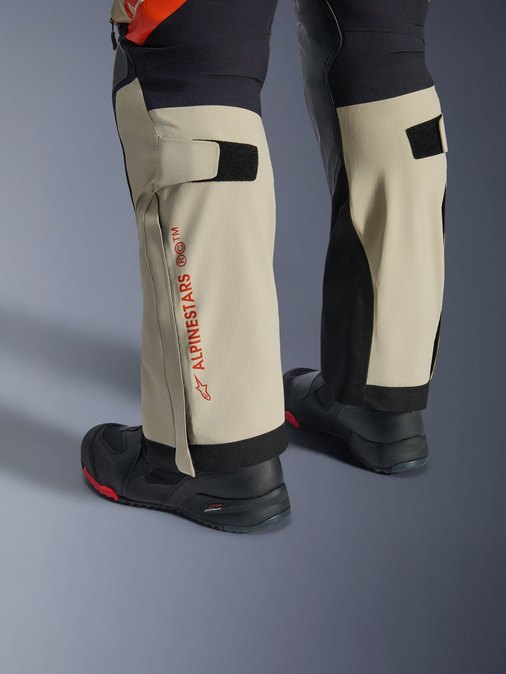 Calças Alpinestars Halo Pro Drystar® XF Laminated, Mastic Black, grande plano da parte inferior das pernas com logótipo Alpinestars vermelho e ajustadores de velcro na panturrilha, usadas com botas de aventura pretas