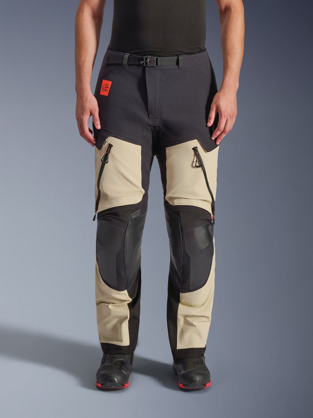 Alpinestars Halo Pro Drystar® XF Laminated Pants, calças de motociclismo de aventura, Mastic Black, vista frontal usada por um piloto, com grandes aberturas com fecho nas coxas, painéis reforçados pretos nos joelhos e construção em têxtil bege, combinadas com botas de motociclismo pretas e vermelhas