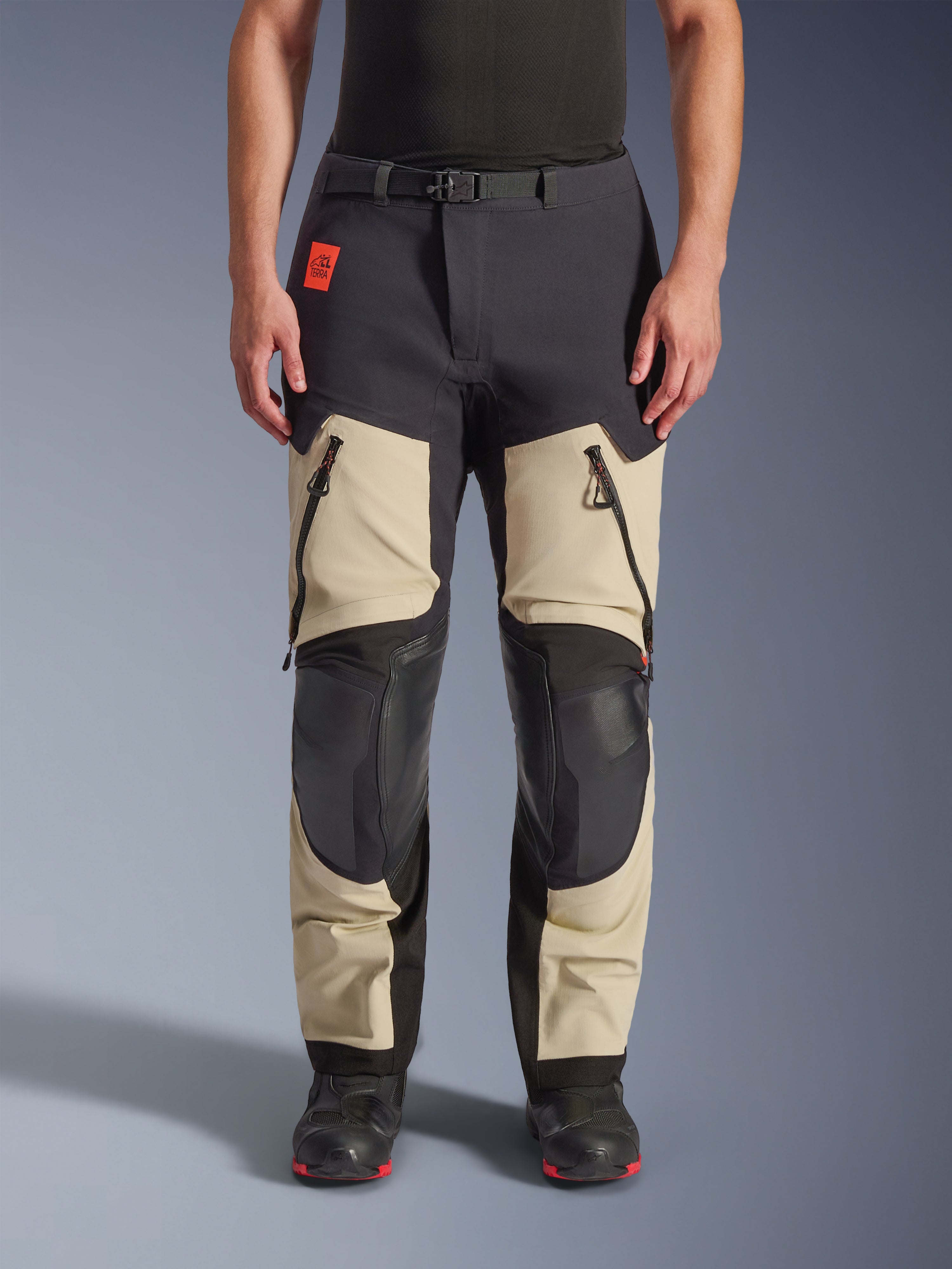 Alpinestars Halo Pro Drystar® XF Laminated Pants, calças de motociclismo de aventura, Mastic Black, vista frontal usada por um piloto, com grandes aberturas com fecho nas coxas, painéis reforçados pretos nos joelhos e construção em têxtil bege, combinadas com botas de motociclismo pretas e vermelhas