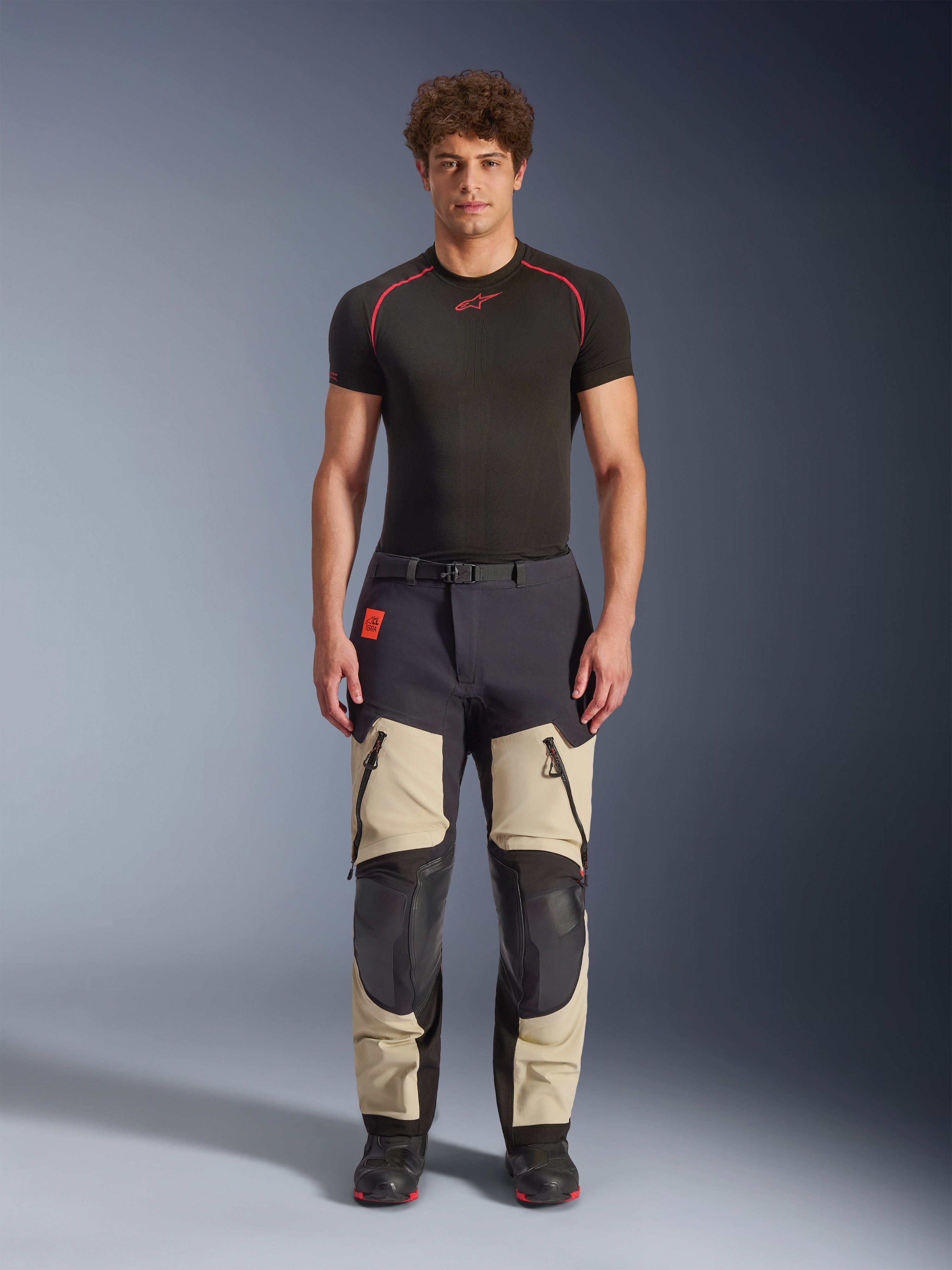 Alpinestars Halo Pro Drystar® XF Laminated Pants, Mastic Black, calças de motociclismo de aventura vestidas por um modelo com uma camada base preta e botas, apresentando painéis bege nas coxas com grandes aberturas de ventilação com fecho, secções reforçadas nos joelhos a preto e um cinto com fivela magnética.