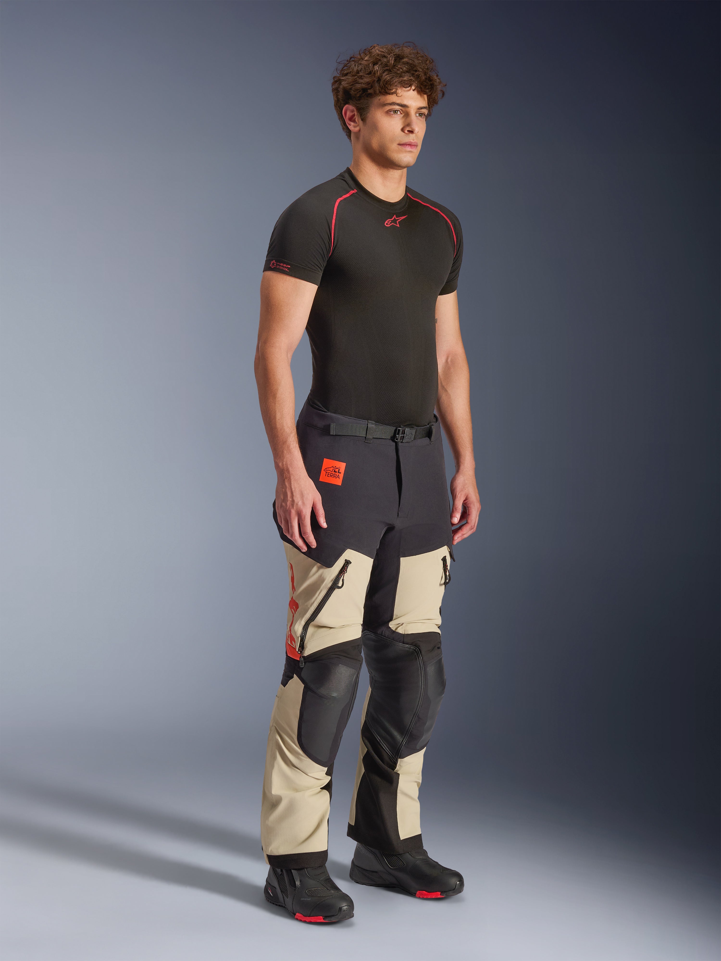 Calças Alpinestars Halo Pro Drystar® XF Laminated em Mastic Black, calças de motociclismo de aventura vestidas por um modelo, com reforços pretos nos joelhos e no assento, logótipo cor-de-laranja e múltiplos fechos de correr funcionais, combinadas com uma camada de base preta e botas de motociclismo.