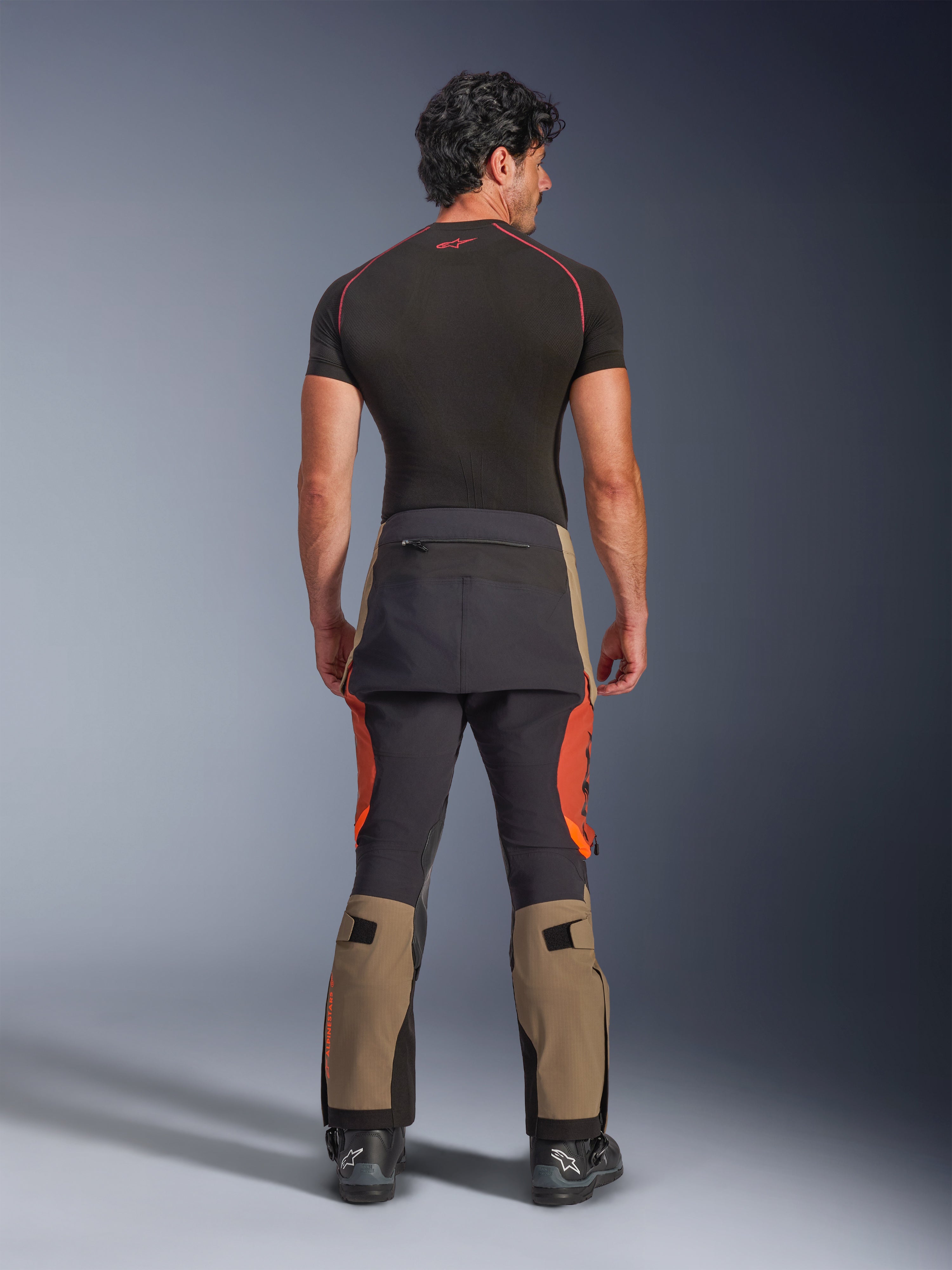 Alpinestars Halo Pro Drystar® XF Laminated Pants, calças de motociclismo de aventura em Walnut Rust Black, vista traseira num modelo a usar botas pretas e uma camada base de manga curta, apresentando a parte inferior das pernas em tom nogueira, detalhes em laranja ferrugem e painéis pretos reforçados.