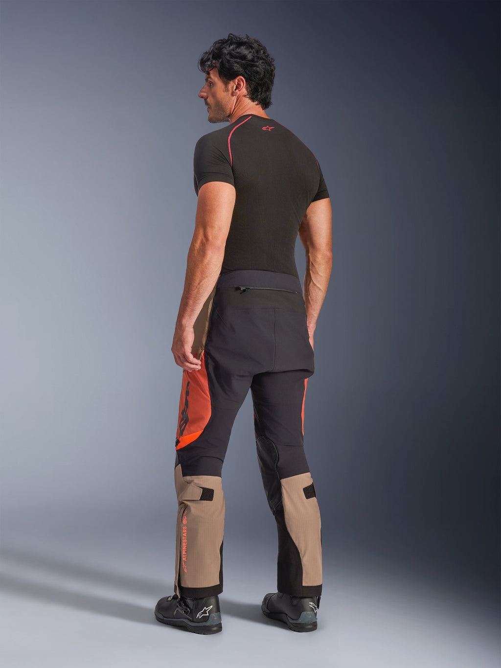 Calças Alpinestars Halo Pro Drystar® XF Laminated em Walnut Rust Black, vista traseira de um modelo masculino a usar calças de motociclismo de aventura com painéis em bege, laranja e preto, combinadas com uma camada base preta de manga curta e botas de motociclismo pretas, destacando o equipamento técnico de condução.