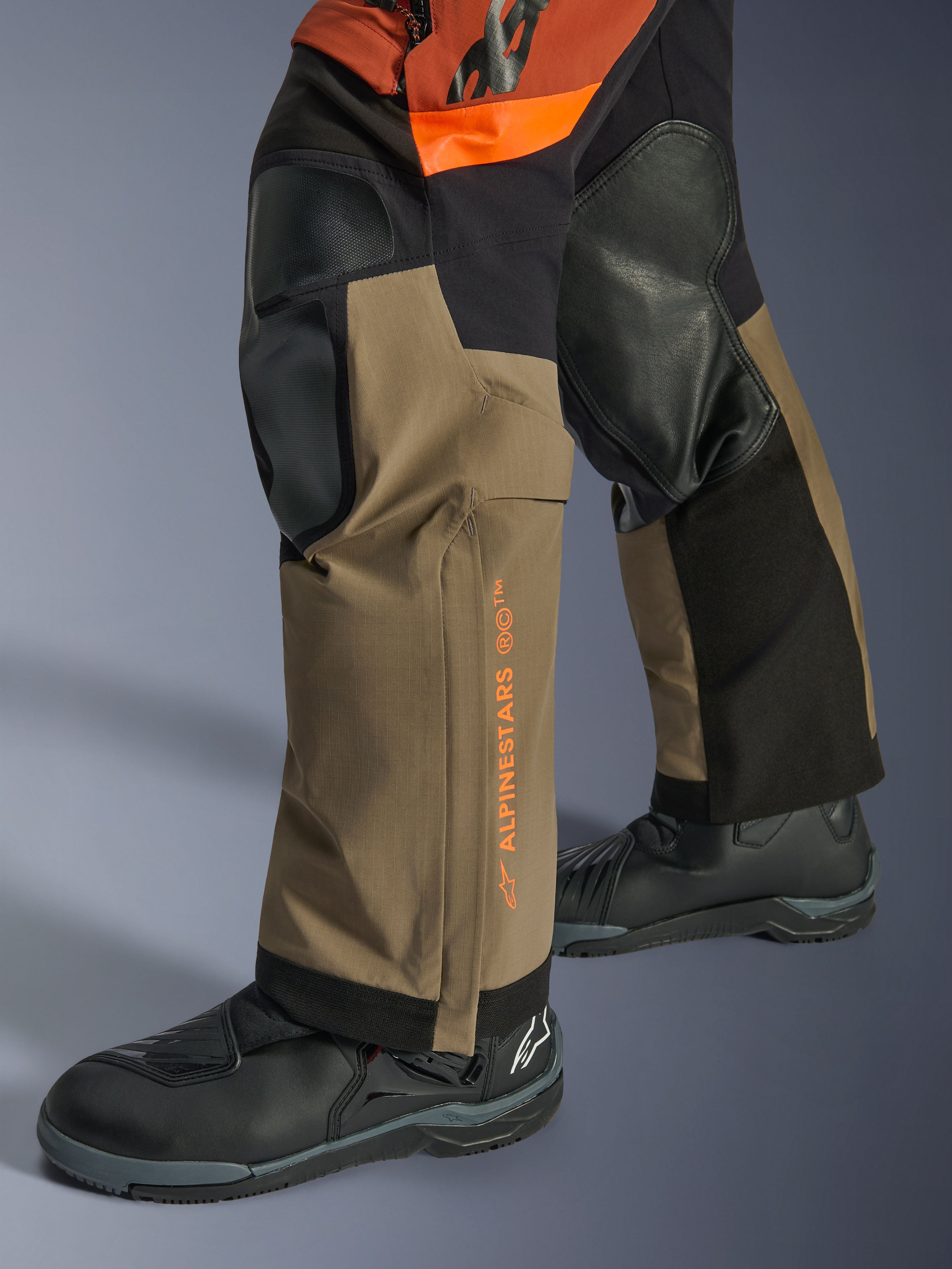 Calças Alpinestars Halo Pro Drystar® XF Laminadas, calças de motociclismo de aventura, Walnut Rust e preto, pormenor com painéis reforçados nos joelhos e marca em cor de laranja, usadas com botas de touring pretas, construção têxtil durável para condução todo-o-terreno