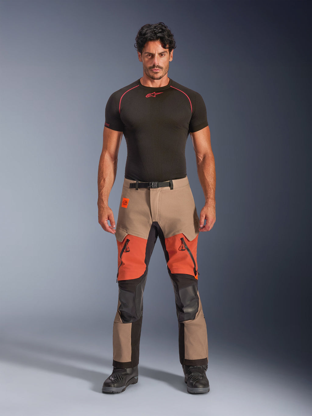 Modelo masculino a usar calças Alpinestars Halo Pro Drystar® XF Laminated, calças de motociclismo de aventura, Walnut Rust Black, design em blocos de cor bege e laranja com painéis técnicos pretos, com aberturas de ventilação com fecho nas coxas e fivela magnética na cintura, combinadas com uma camada base preta e botas de motociclismo pretas