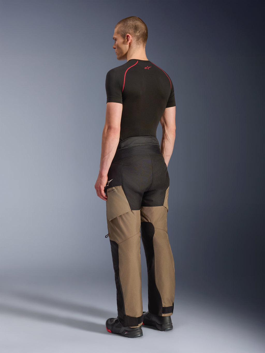 Calças Alpinestars Andes V4 Drystar®, calças de motociclismo de aventura, Walnut Black, vista traseira de três quartos no modelo, com painéis reforçados em nogueira e preto, usadas com uma camada base preta e botas de motociclismo