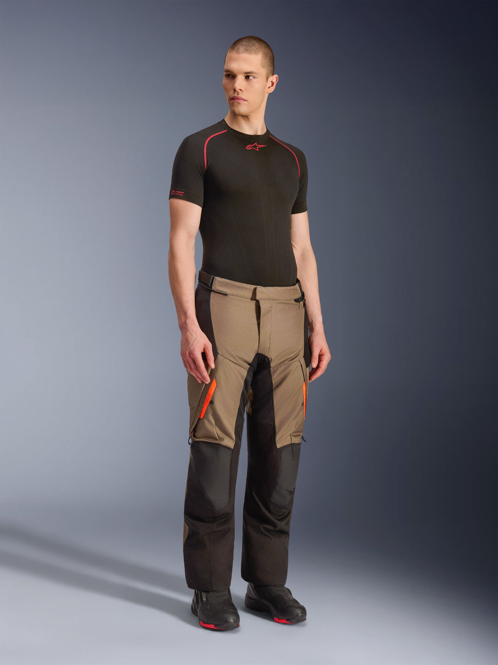 Calças Alpinestars Andes V4 Drystar®, calças de motociclismo adventure touring, cor Walnut Black com detalhes laranja, vestidas por modelo com camada de base preta de manga curta e botas pretas, com painéis têxteis reforçados e bolsos cargo, design robusto para condução em todas as condições climatéricas