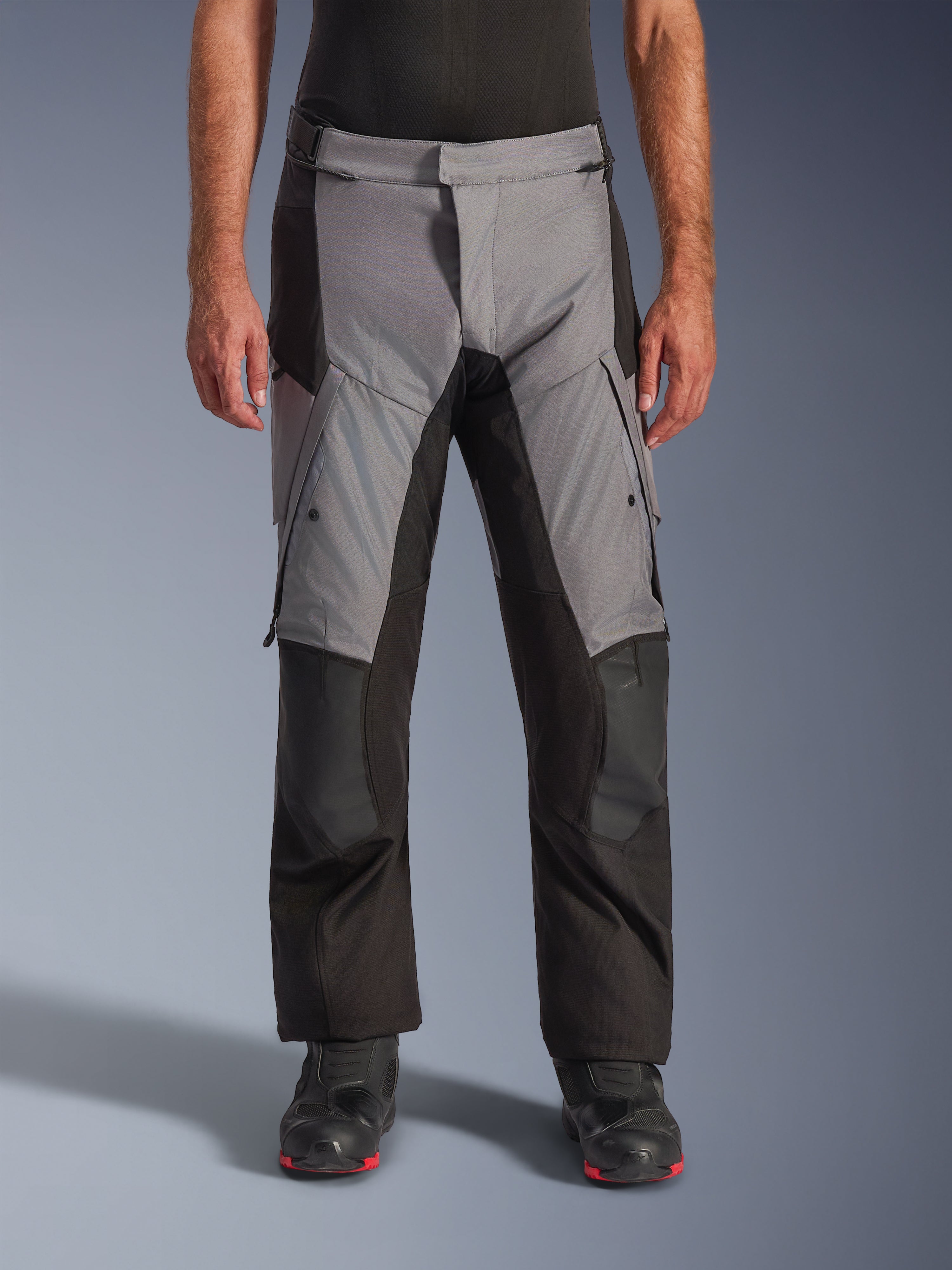 Alpinestars Andes V4 Drystar® Pants, calças de motociclismo para turismo e aventura, cinzentas e pretas, vista frontal vestidas por um modelo com botas de motociclista pretas, com reforços nos joelhos e bolsos cargo nas coxas, construção têxtil impermeável e respirável para condução em todas as condições meteorológicas