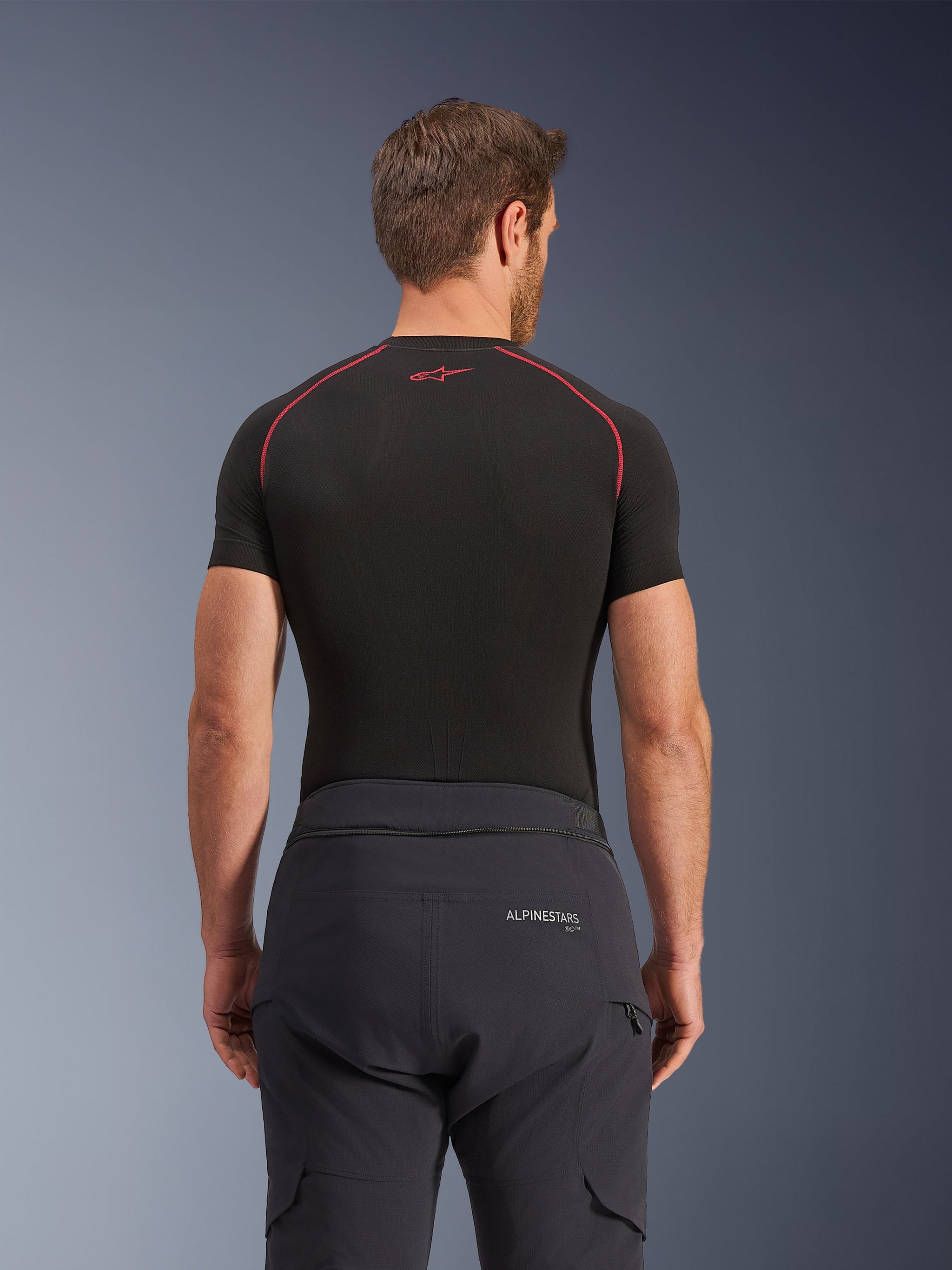 Calças Alpinestars Andes Pro Drystar® XF, calças de adventure touring, pretas e cinzento escuro, vista traseira mostrando a marca na cintura e o fecho de ligação ao casaco, usadas com uma camada de base de manga curta preta com costuras vermelhas