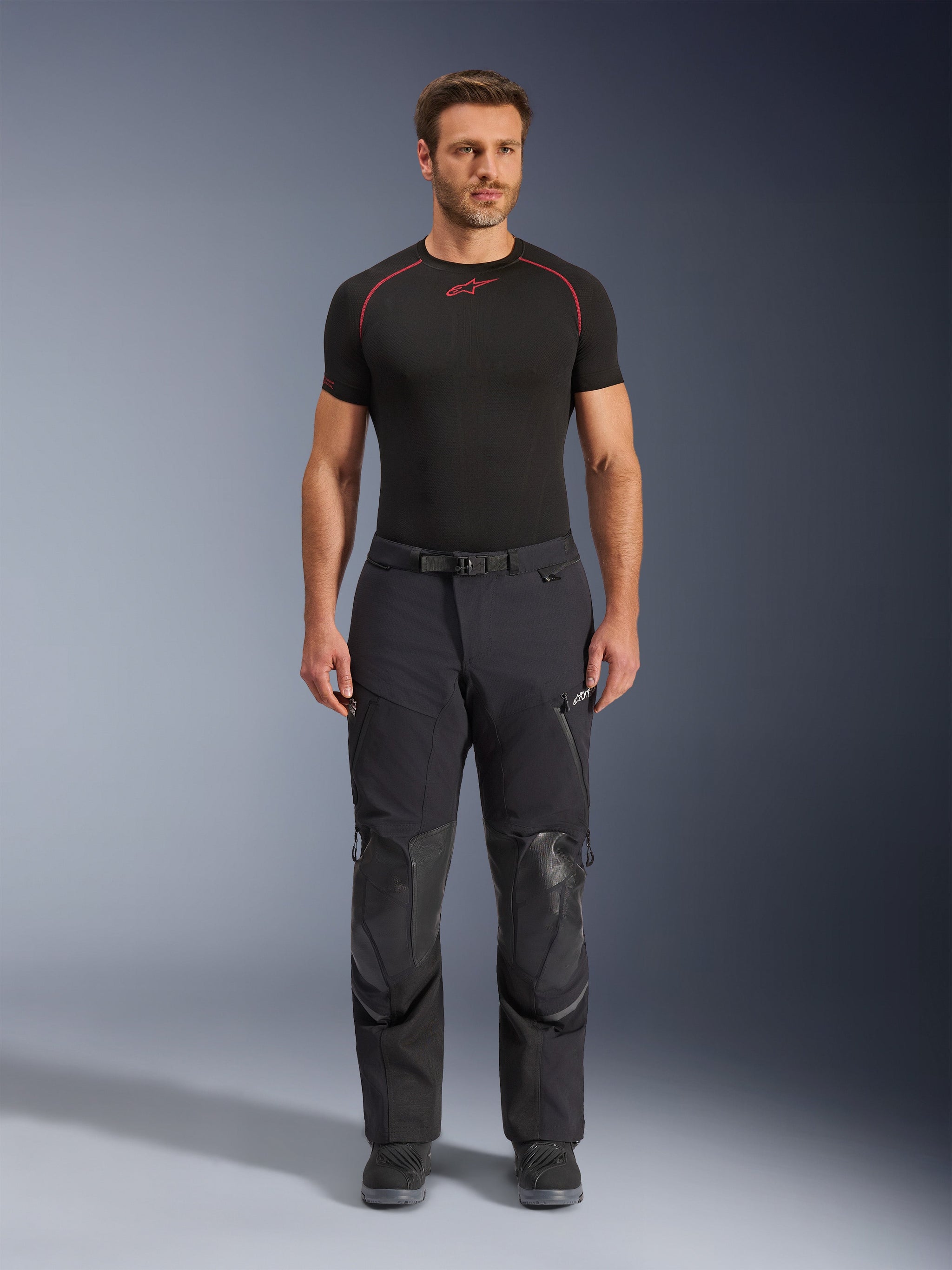 Alpinestars Andes Pro Drystar® XF Pants, pretas, calças de motociclismo de aventura vestidas por um modelo numa vista frontal, com painéis de reforço em pele nos joelhos e fivela magnética na cintura, combinadas com uma t-shirt de camada base Alpinestars preta com detalhes vermelhos
