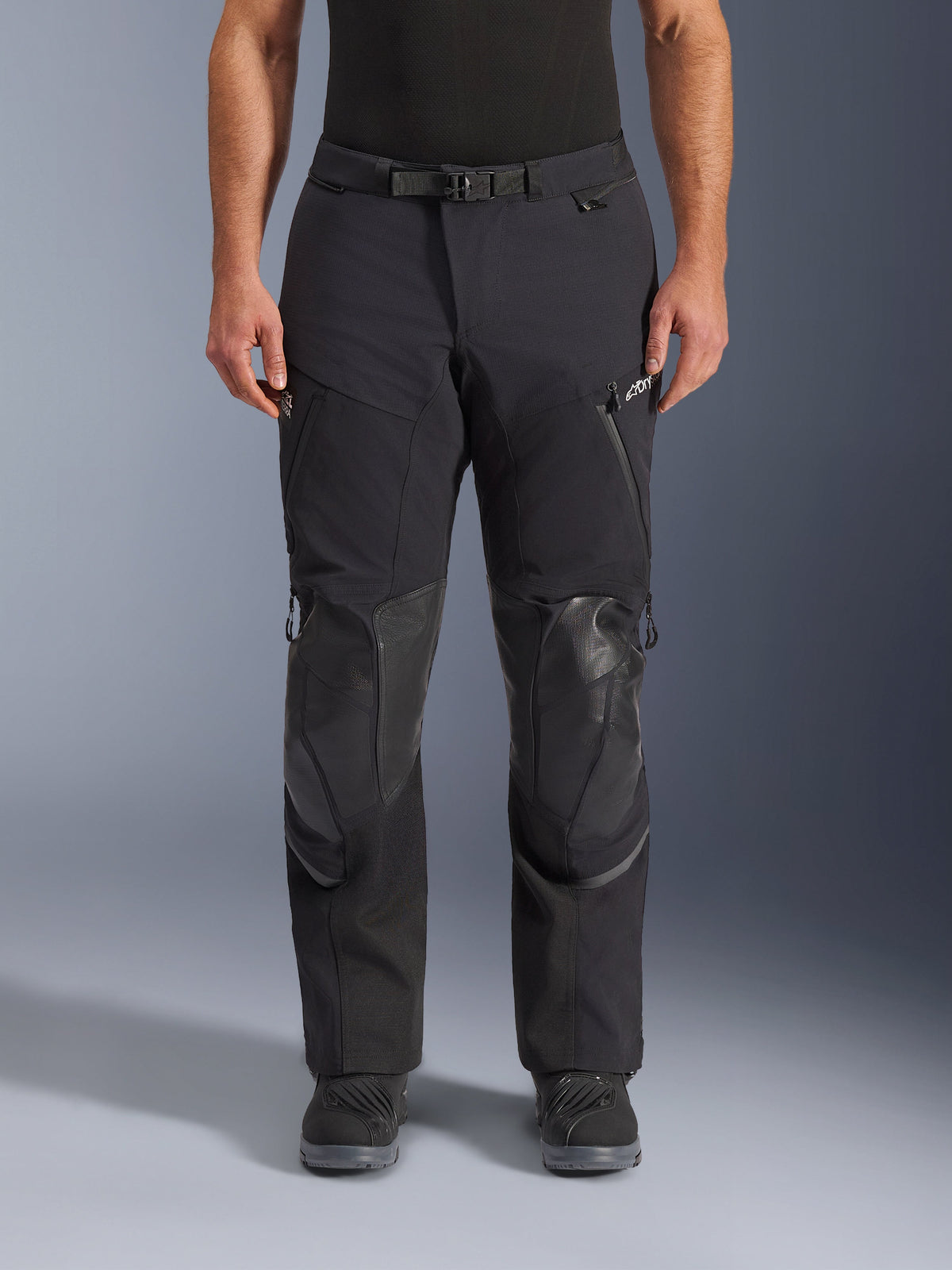 Alpinestars Andes Pro Drystar® XF Pants, calças de motociclismo adventure touring, em preto e cinzento escuro, vista frontal, com construção laminada de 3 camadas, painéis de proteção de joelho integrados, bolsos com fecho impermeável e fivela de cintura magnética, combinadas com botas de motociclismo pretas.