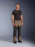 Calças Alpinestars Cusco Drystar®, calças de motociclismo de aventura, em nogueira e preto, usadas por um modelo com uma base layer preta e botas, com bolsos cargo grandes nas coxas, fechos de ventilação e um design robusto em dois tons