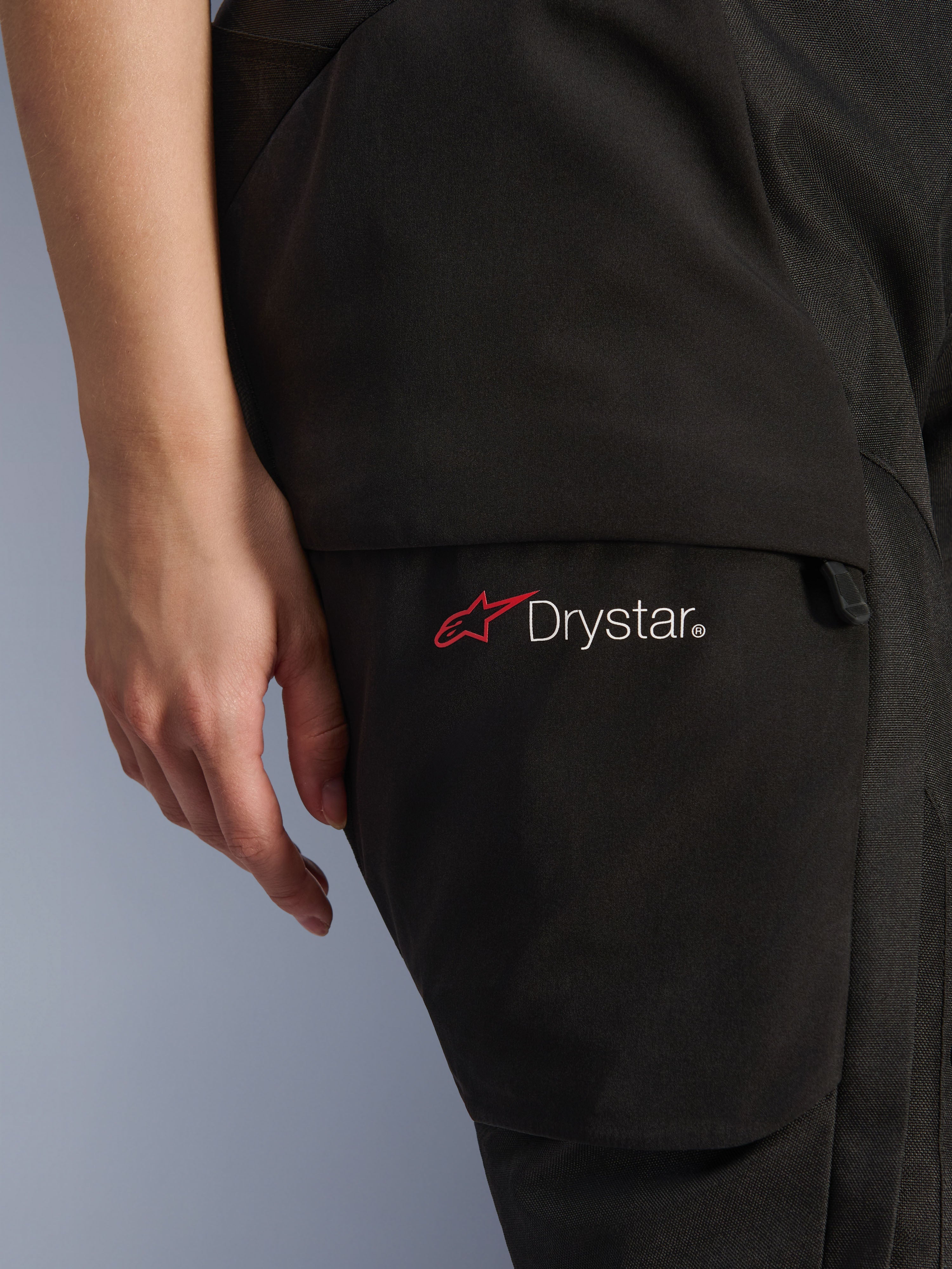 Calças Alpinestars Stella Andes V4 Drystar®, calças de motociclismo de aventura, pretas, primeiro plano da zona da coxa com o logótipo Drystar e o ícone da estrela vermelha, material têxtil resistente com bolso cargo e detalhe de fecho de correr