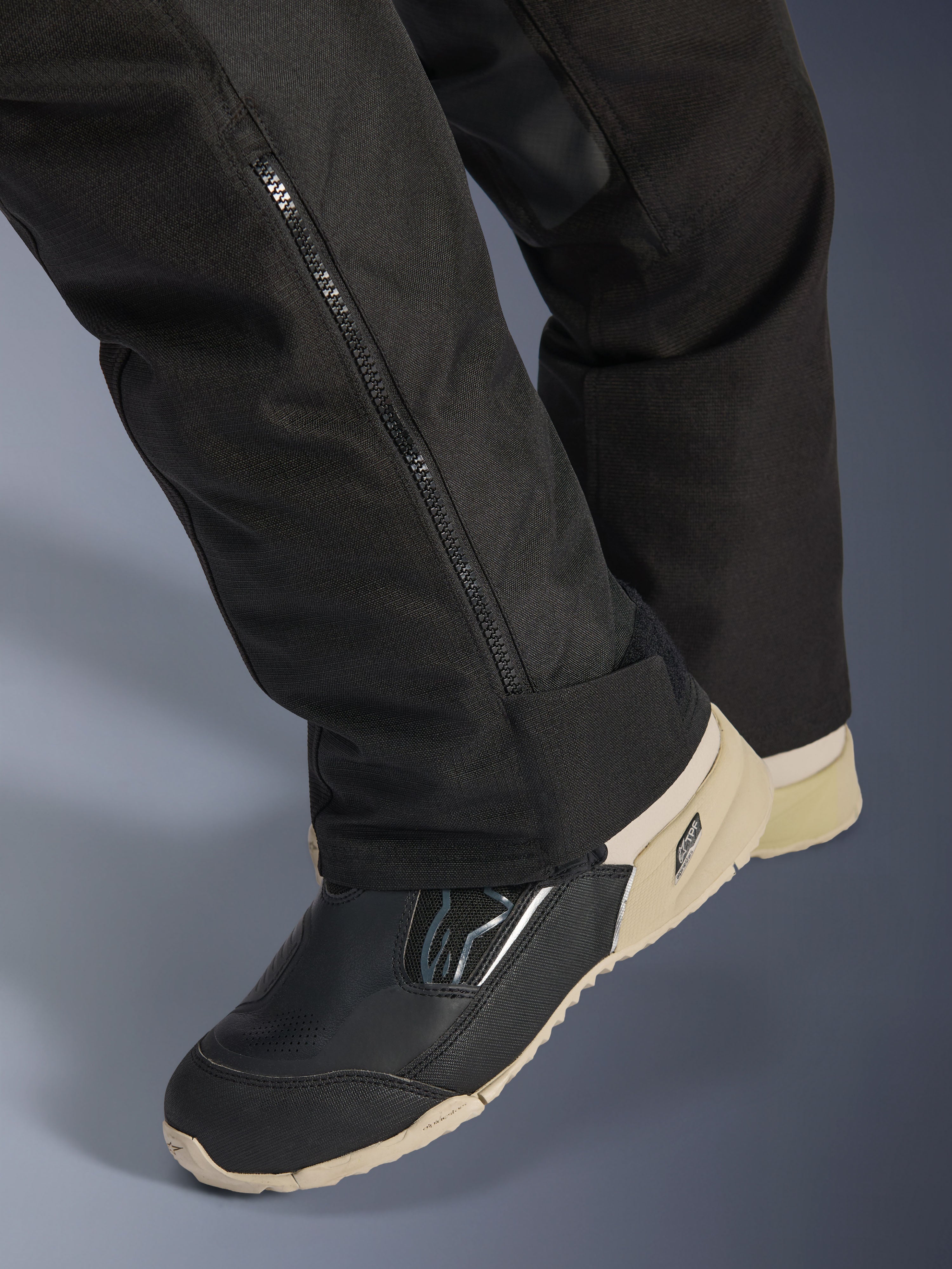 Calças Alpinestars Stella Andes V4 Drystar®, pretas, pormenor da parte inferior da perna com fecho lateral para ajuste da bota, material têxtil durável, usadas com botas de motociclismo pretas e beges, design funcional pronto para a aventura