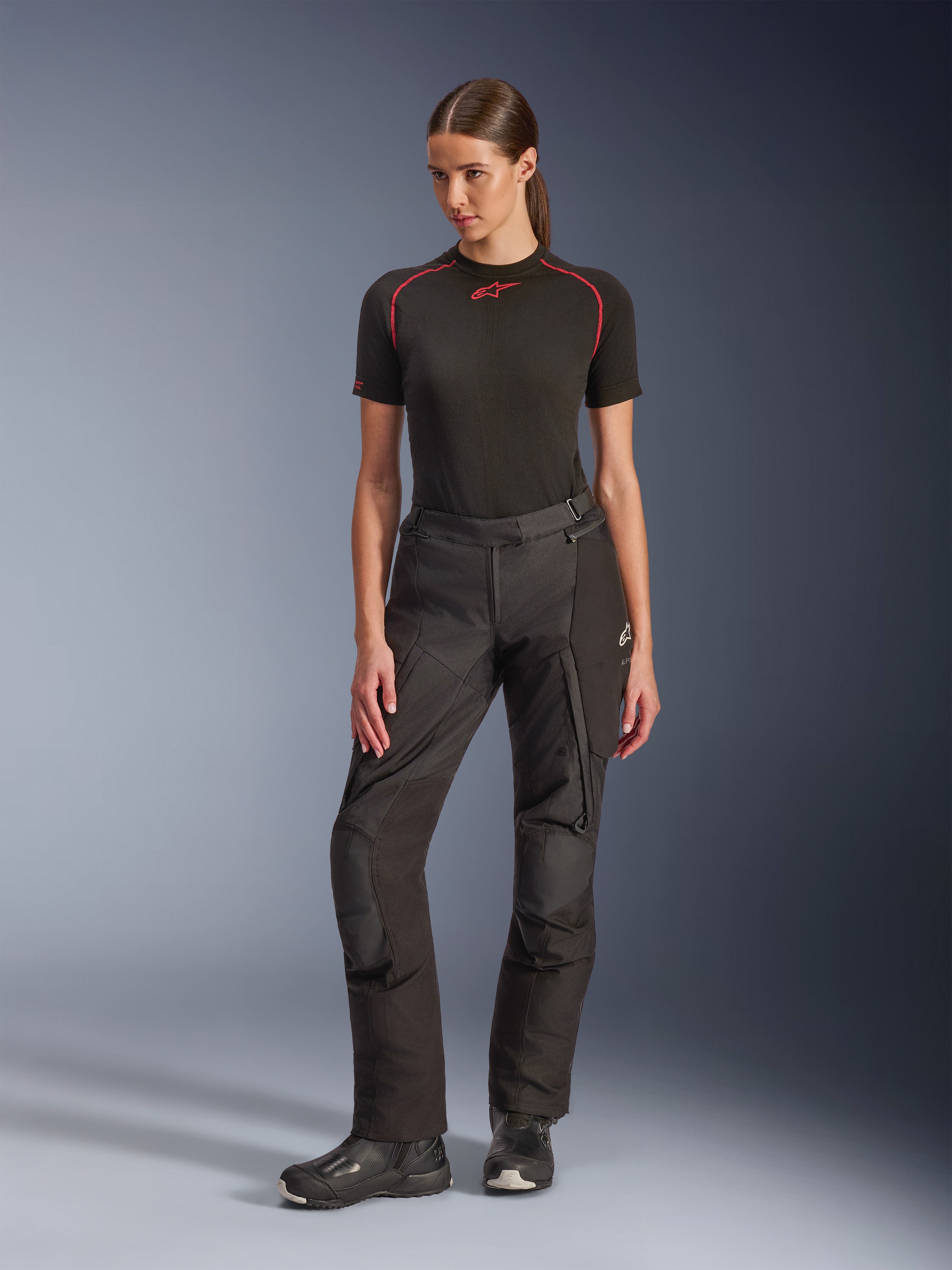 Calças Alpinestars Stella Andes V4 Drystar®, calças de aventura de motociclismo para senhora, pretas, vestidas por uma modelo com uma camada de base preta de manga curta e botas de motociclismo pretas, com joelhos reforçados e bolsos cargo, design ergonómico para viagens em todas as condições meteorológicas