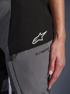 Pormenor das calças Alpinestars Stella Andes V4 Drystar® para mulher em preto e cinzento escuro, com logótipo de estrela branca num painel preto e marca Alpinestars a preto num painel cinzento, mostrando o tecido técnico e o detalhe do bolso com fecho de correr.
