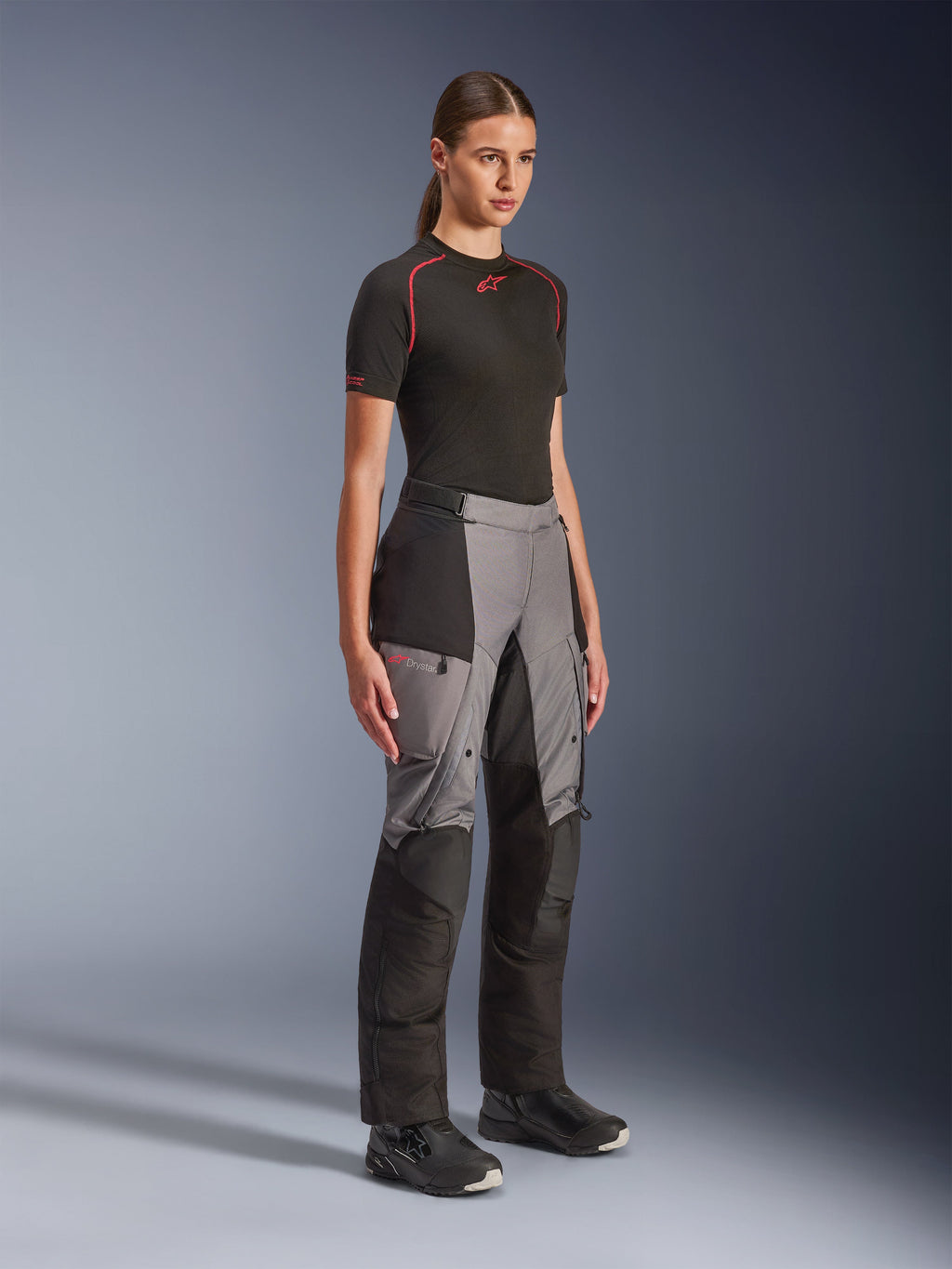 Calças Alpinestars Stella Andes V4 Drystar®, calças de motociclismo de aventura para mulher, pretas e cinzento escuro, vestidas por uma modelo com uma base layer Alpinestars preta e botas pretas, com painéis reforçados, logótipo Drystar vermelho e bolsos cargo.