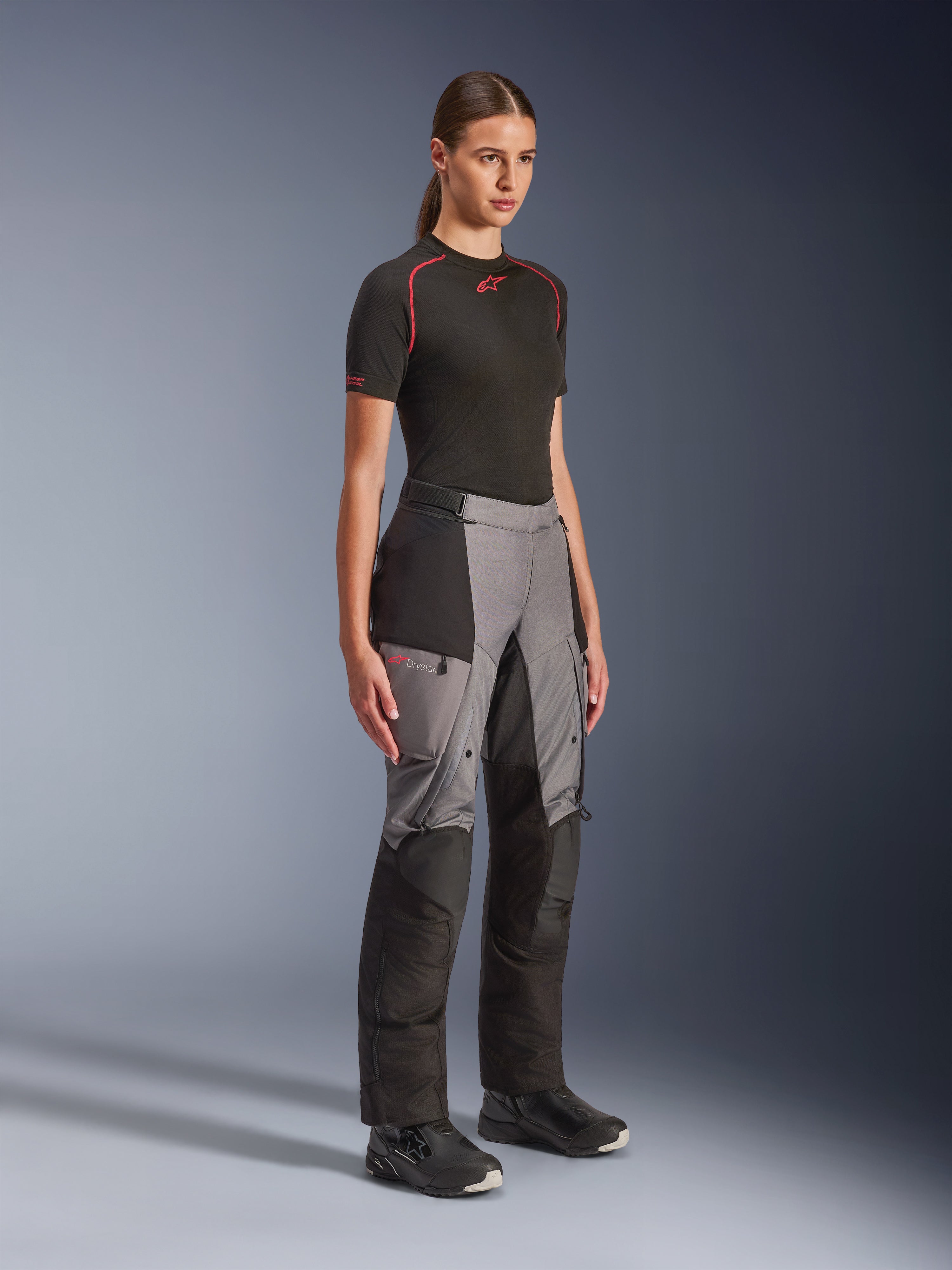 Calças Alpinestars Stella Andes V4 Drystar®, calças de motociclismo de aventura para mulher, pretas e cinzento escuro, vestidas por uma modelo com uma base layer Alpinestars preta e botas pretas, com painéis reforçados, logótipo Drystar vermelho e bolsos cargo.