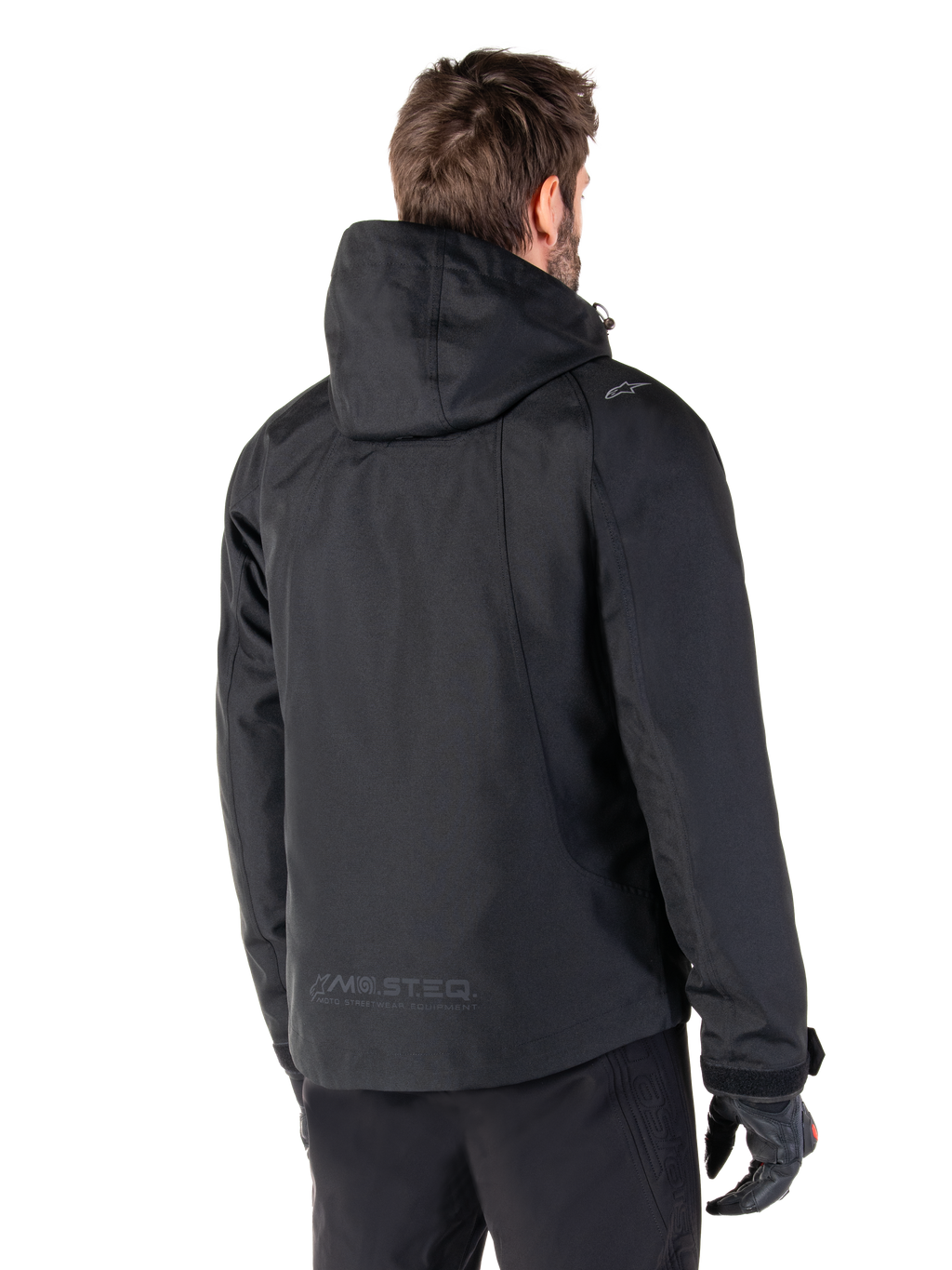 Alpinestars Morush Waterproof Parka, Black, rear view of a man wearing the hooded motorcycle jacket, featuring raglan sleeves, an extended rear hem for coverage, MO.ST.EQ. branding on the lower back, and a subtle Alpinestars logo on the shoulder, urban streetwear style