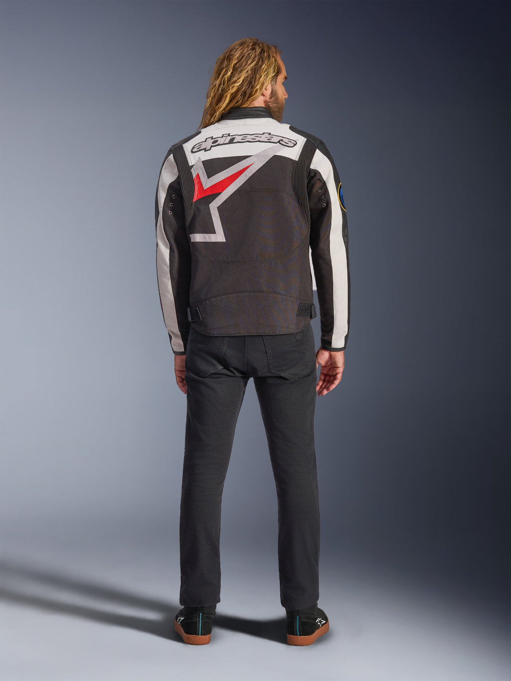 Casaco Alpinestars Axton, casaco de motociclismo, preto, branco sujo e vermelho vivo, vista traseira num modelo, com um logótipo de estrela Alpinestars de grandes dimensões nas costas, painéis das mangas em branco sujo, usado com calças pretas, design inspirado no vestuário de equipa de corrida vintage