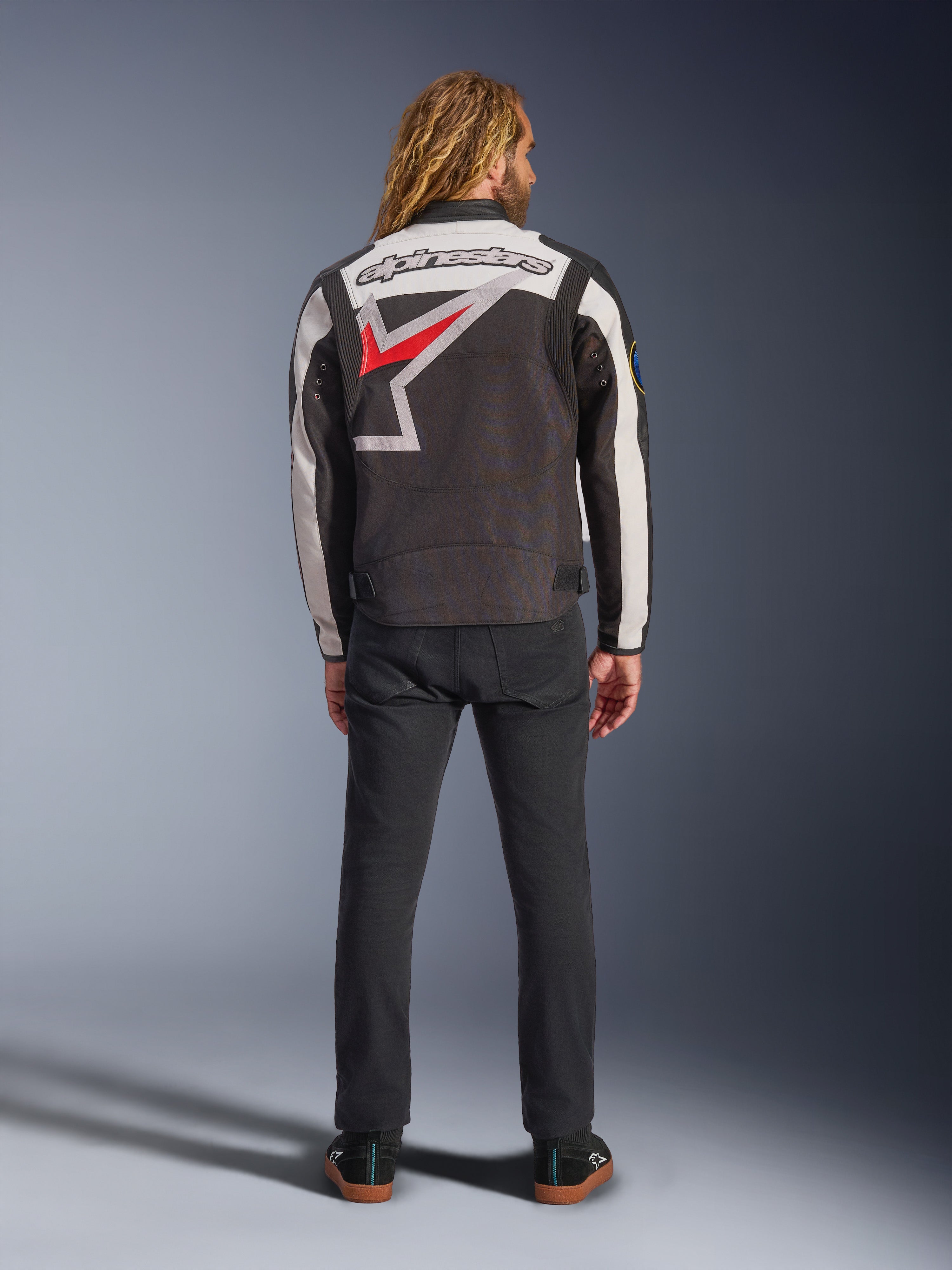 Casaco Alpinestars Axton, casaco de motociclismo, preto, branco sujo e vermelho vivo, vista traseira num modelo, com um logótipo de estrela Alpinestars de grandes dimensões nas costas, painéis das mangas em branco sujo, usado com calças pretas, design inspirado no vestuário de equipa de corrida vintage