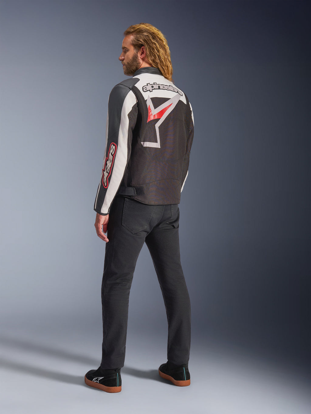 Casaco Alpinestars Axton, casaco de motociclismo, preto, branco sujo e vermelho vivo, vista traseira usada por modelo masculino, com grande logótipo de estrela e marca Alpinestars nas costas, design de inspiração vintage com painéis de acordeão, combinado com calças pretas e sapatilhas