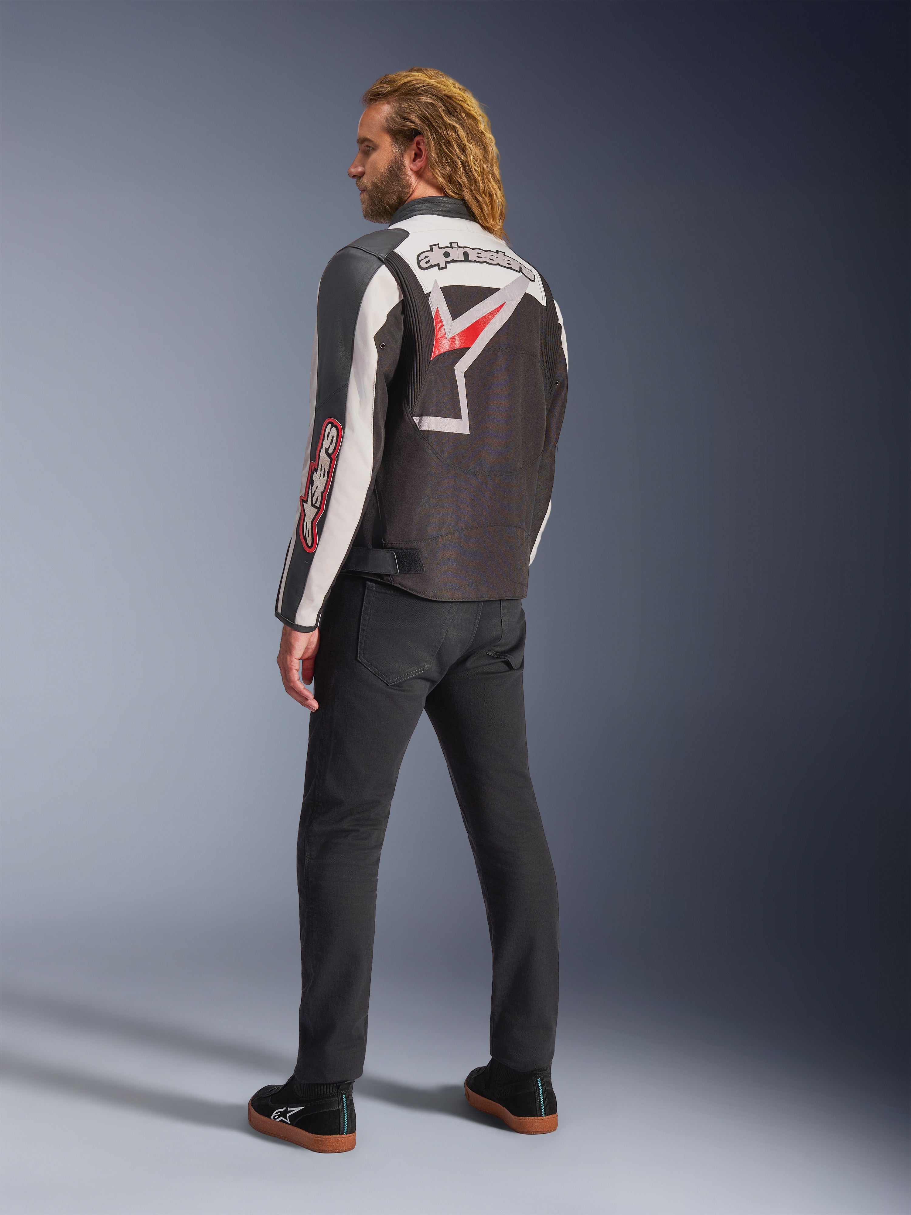 Casaco Alpinestars Axton, casaco de motociclismo, preto, branco sujo e vermelho vivo, vista traseira usada por modelo masculino, com grande logótipo de estrela e marca Alpinestars nas costas, design de inspiração vintage com painéis de acordeão, combinado com calças pretas e sapatilhas
