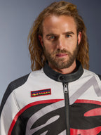 Homem a usar o Casaco Alpinestars Axton, casaco de motociclismo de inspiração vintage em Preto, Off White e Vermelho Brilhante, vista em grande plano do emblema da marca no peito e gola de motociclista minimalista, equipamento contemporâneo para condução em estrada