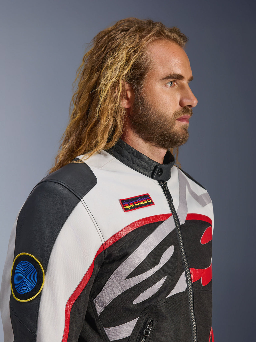 Homem com cabelo comprido e barba a usar o Casaco Alpinestars Axton, casaco de motociclismo, preto, off-white e vermelho vivo, design inspirado em corridas vintage com remendos da marca e logótipo envolvente, vista de perfil em grande plano, estilo urbano contemporâneo