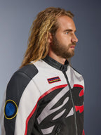Homem com cabelo comprido e barba a usar o Casaco Alpinestars Axton, casaco de motociclismo, preto, off-white e vermelho vivo, design inspirado em corridas vintage com remendos da marca e logótipo envolvente, vista de perfil em grande plano, estilo urbano contemporâneo