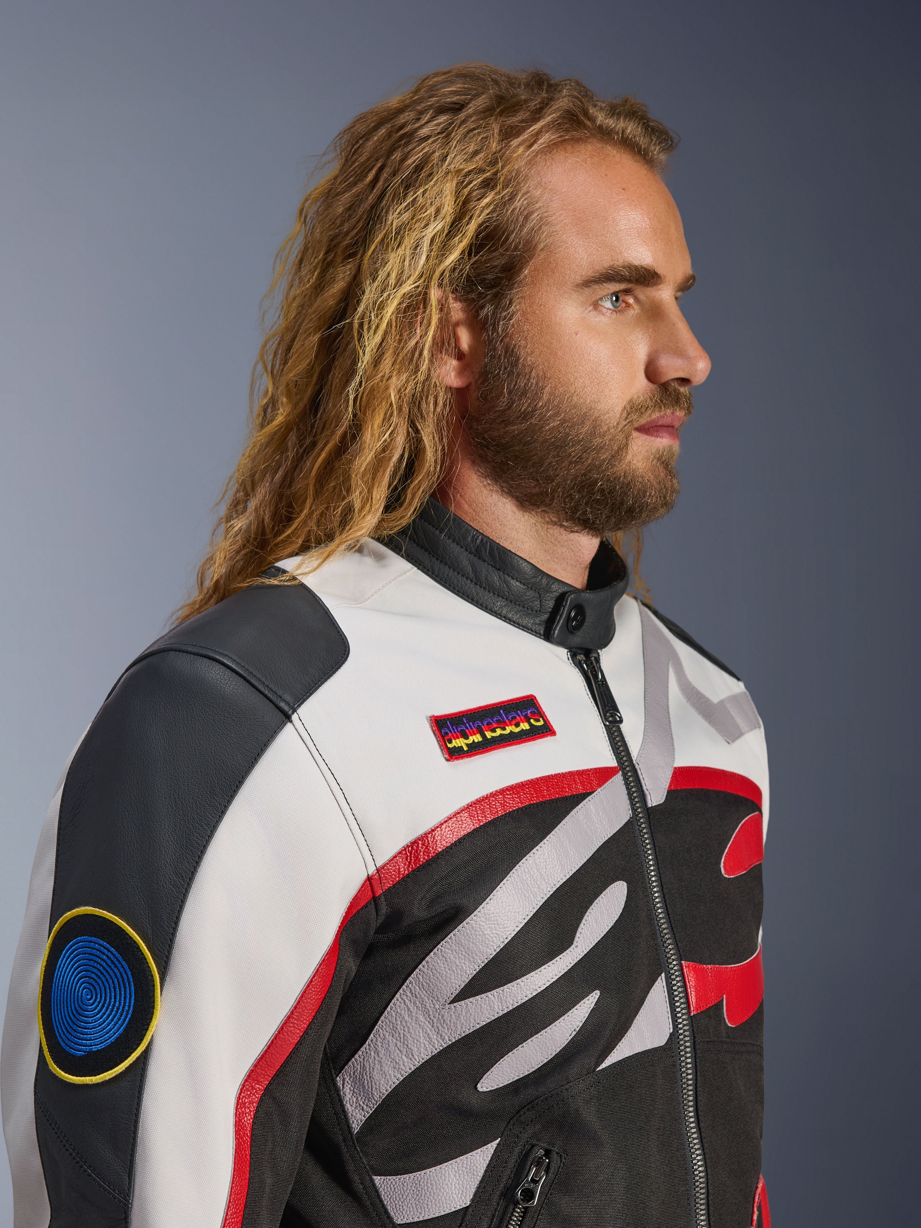 Homem com cabelo comprido e barba a usar o Casaco Alpinestars Axton, casaco de motociclismo, preto, off-white e vermelho vivo, design inspirado em corridas vintage com remendos da marca e logótipo envolvente, vista de perfil em grande plano, estilo urbano contemporâneo
