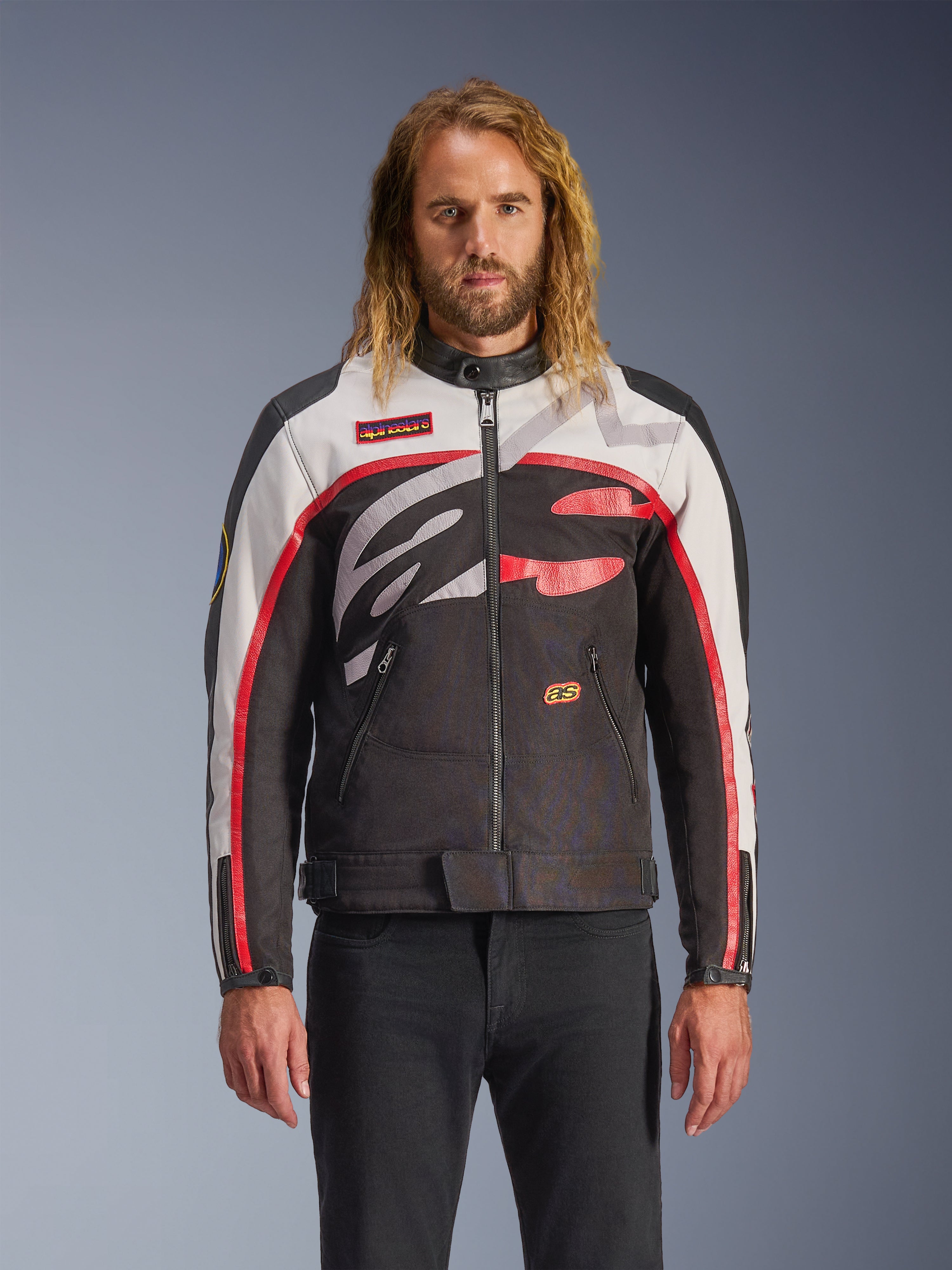 Casaco Alpinestars Axton, casaco de motociclismo, preto, branco e vermelho vivo, com padrões abstratos no peito e logótipo envolvente, mangas com riscas vermelhas, design de inspiração vintage e gola minimalista, usado por modelo com calças de ganga pretas
