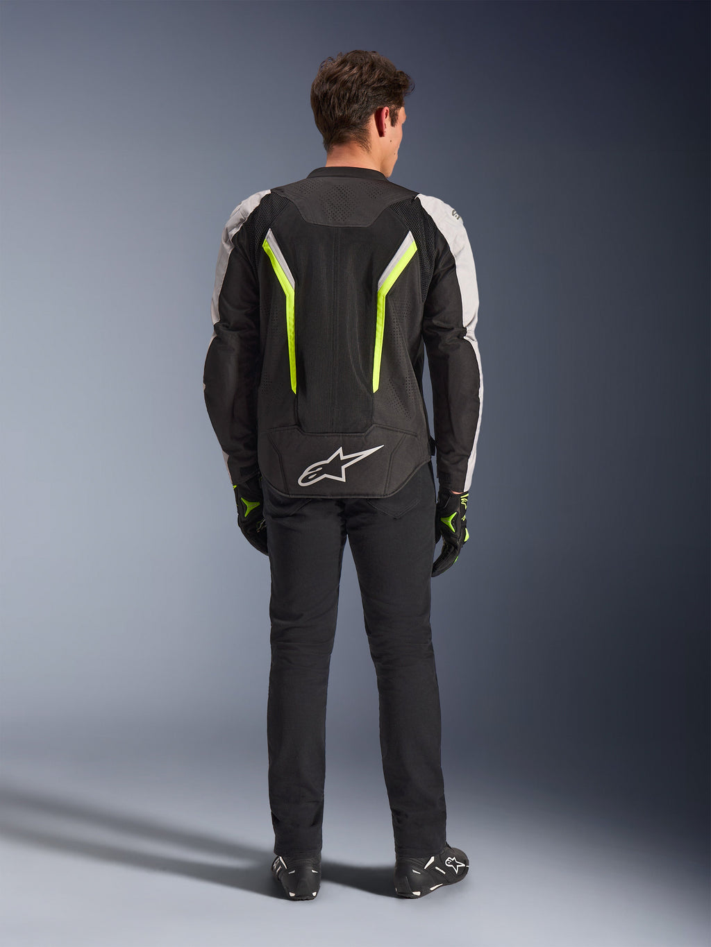 Alpinestars AST V3 Air Jacket, textile motorcycle jacket, Black Silver and Yellow Fluo, back view showing mesh ventilation panels and fluorescent accents, worn with black pants and riding gloves, sport performance riding gear