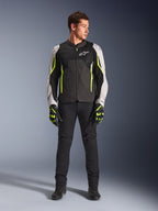 Male model wearing Alpinestars AST V3 Air Jacket in Black Silver Yellow Fluo, featuring mesh ventilation panels and fluorescent accents, paired with black riding pants and matching motorcycle gloves, sport-riding performance gear.