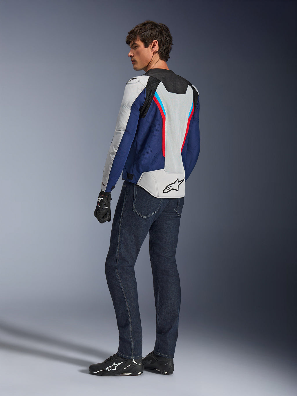 Casaco Alpinestars AST V3 Air, casaco de motociclismo em prateado, azul escuro e vermelho vivo, vista traseira, usado com calças de ganga de motociclismo azul escuro e equipamento preto, com painéis de malha respirável e logótipo Alpinestars nas costas, equipamento técnico de condução com corte desportivo.