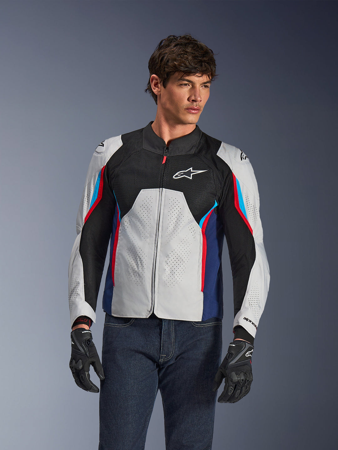 Casaco Alpinestars AST V3 Air em Prateado, Azul Escuro e Vermelho Vivo, vestido por um modelo com calças de ganga escuras e luvas pretas, com painéis prateados perfurados, ventilação em malha preta e detalhes desportivos em vermelho e azul.