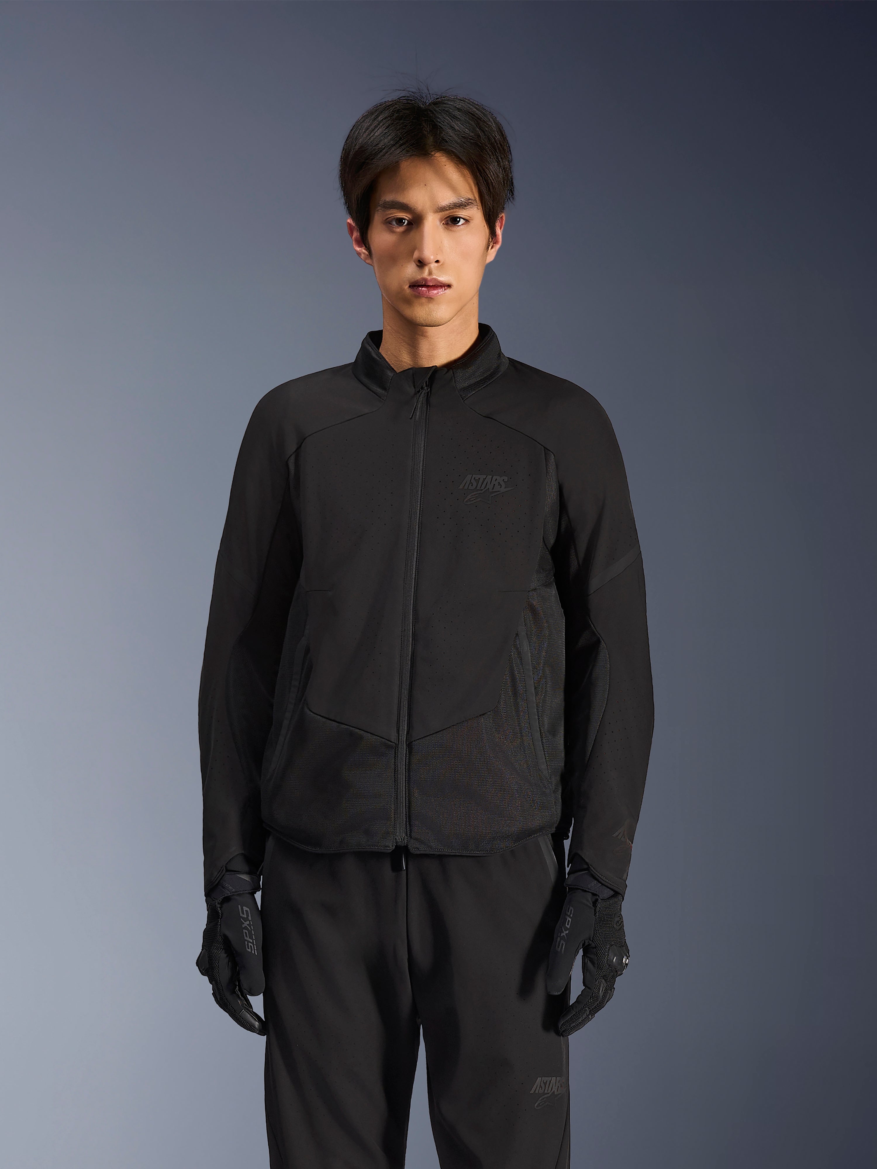 Casaco Alpinestars Aeroshell Airflow em Black Anthracite, casaco de motociclismo leve com ventilação perfurada a laser no peito e nas mangas, painéis de malha respirável e um design elegante de corte desportivo, usado por um modelo com luvas de condução pretas.