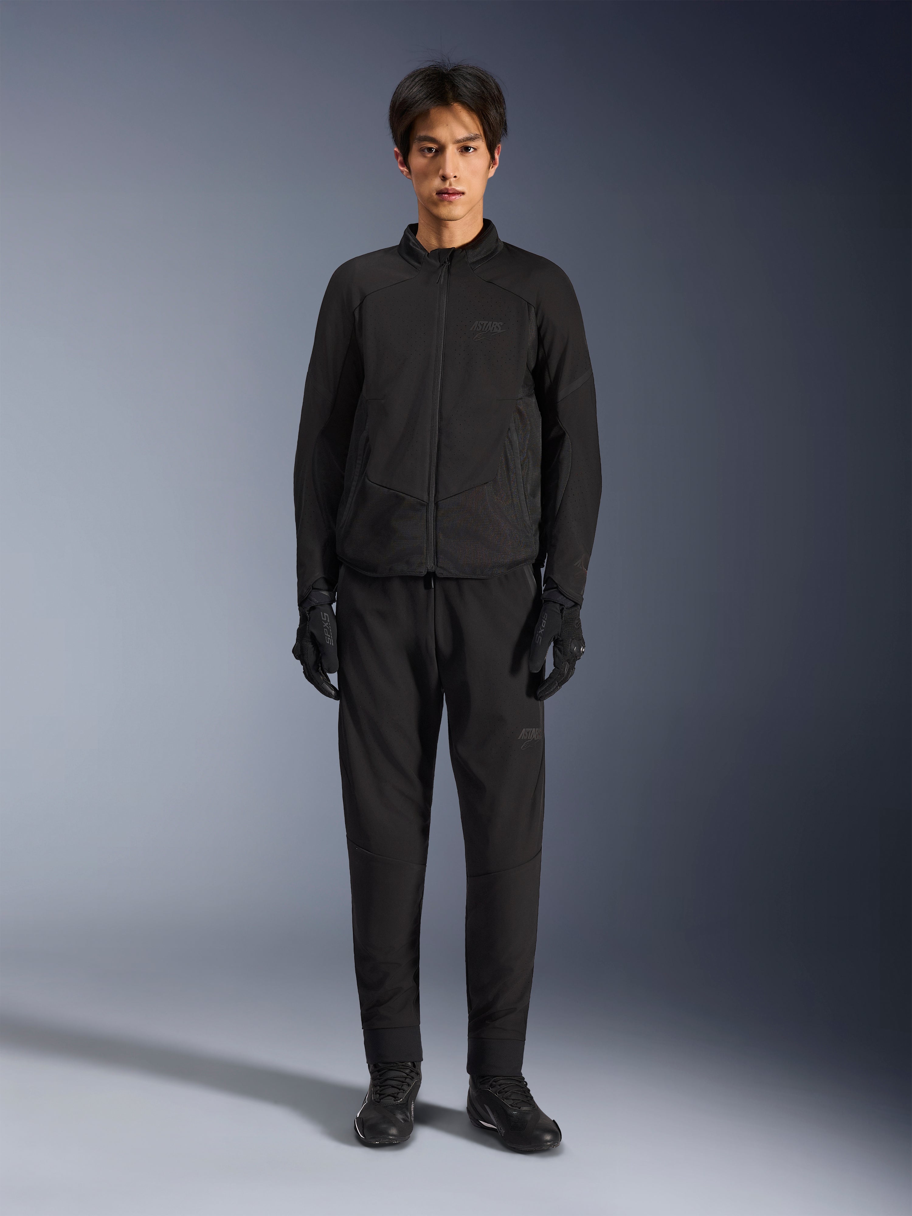 Casaco Alpinestars Aeroshell Airflow, casaco de motociclismo preto, usado por modelo com calças e luvas pretas a condizer, com peito perfurado a laser e painéis de malha laterais, corte desportivo para desempenho em clima quente