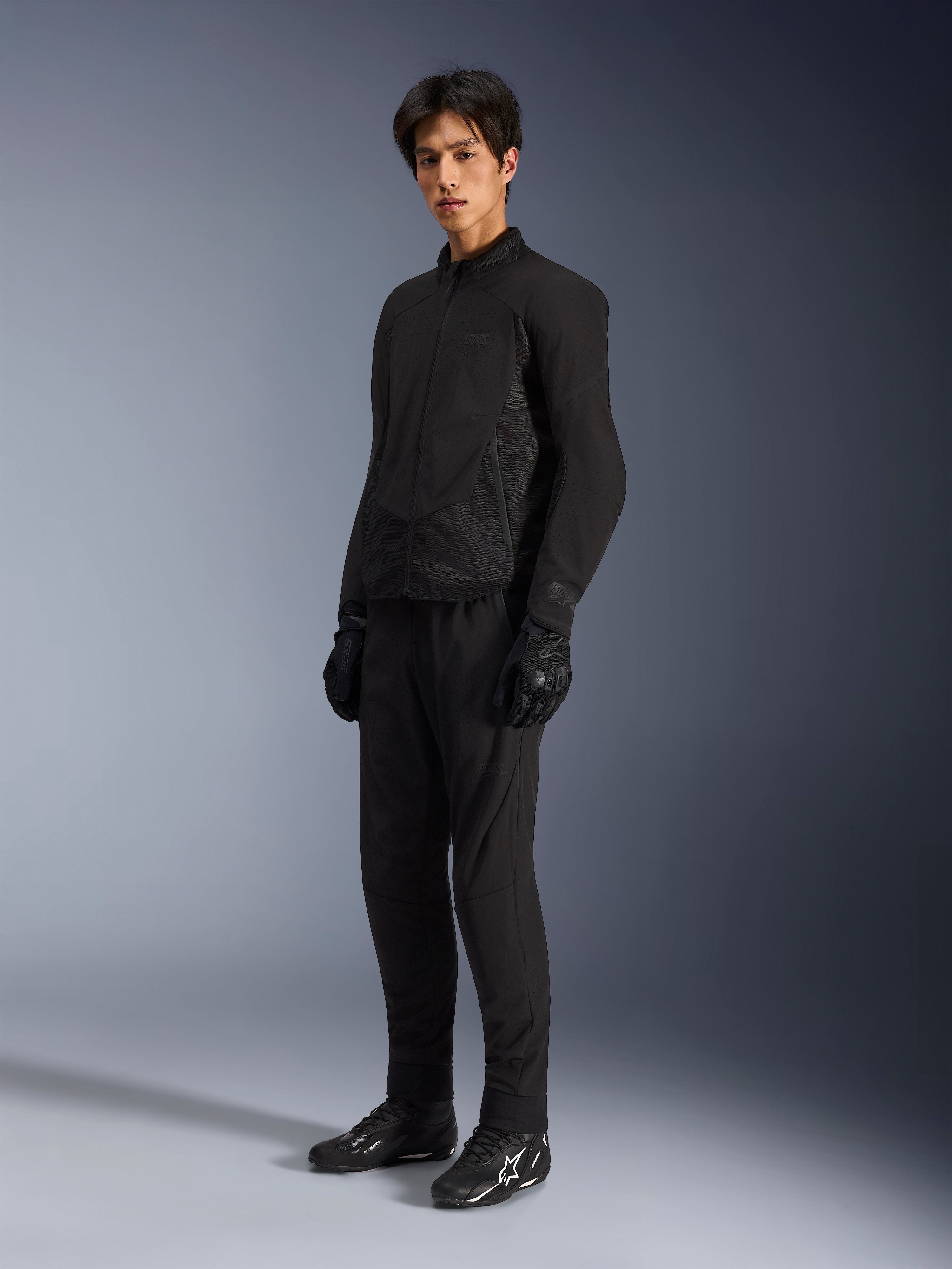 Casaco Alpinestars Aeroshell Airflow, casaco de motociclismo Preto Antracite, com ventilação perfurada a laser e painéis de malha para fluxo de ar, usado por um modelo masculino com calças de condução pretas a condizer, luvas e botas pretas com logótipos brancos, design de corte desportivo para desempenho em estrada