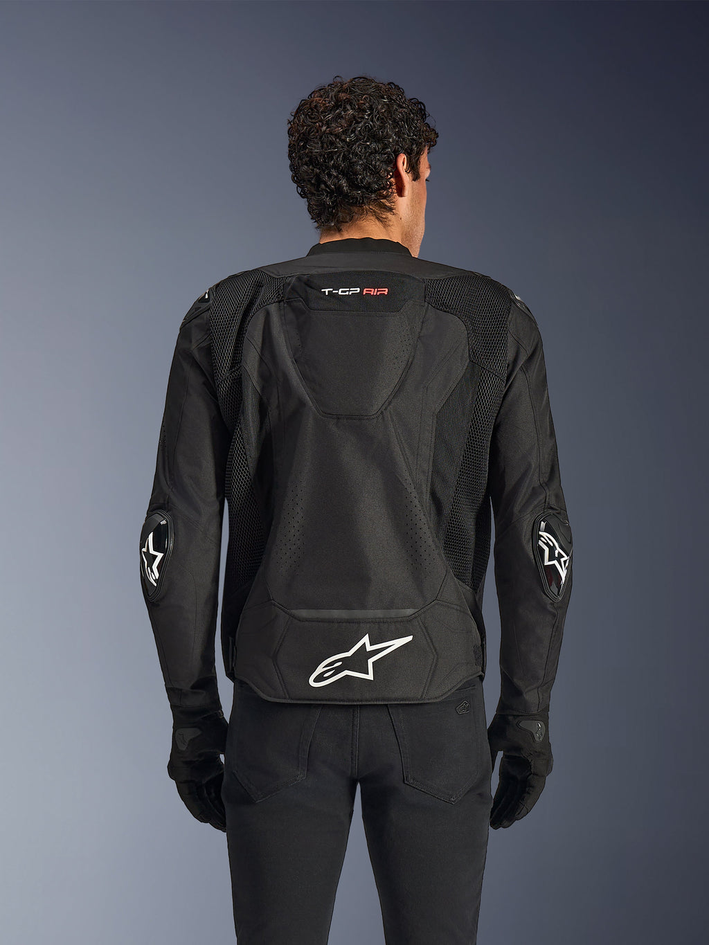 Casaco Alpinestars T-GP Air, Preto, vista traseira de casaco de motociclismo têxtil, com uma corcunda aerodinâmica, grandes painéis de ventilação em malha e logótipo Alpinestars branco na parte inferior das costas, combinado com luvas e calças de motociclismo pretas
