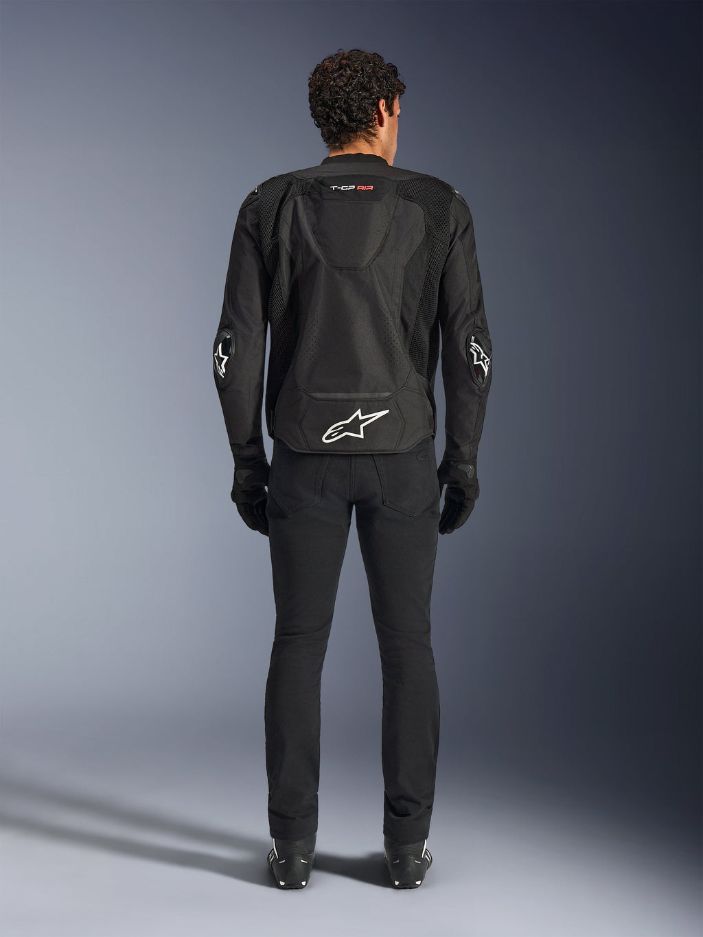Casaco Alpinestars T-GP Air, Preto, vista traseira de um homem a usar o casaco de motociclista têxtil, com painéis de ventilação em malha, uma corcunda aerodinâmica e logótipos Alpinestars brancos na zona lombar e nos cotovelos, usado com calças de condução e luvas pretas