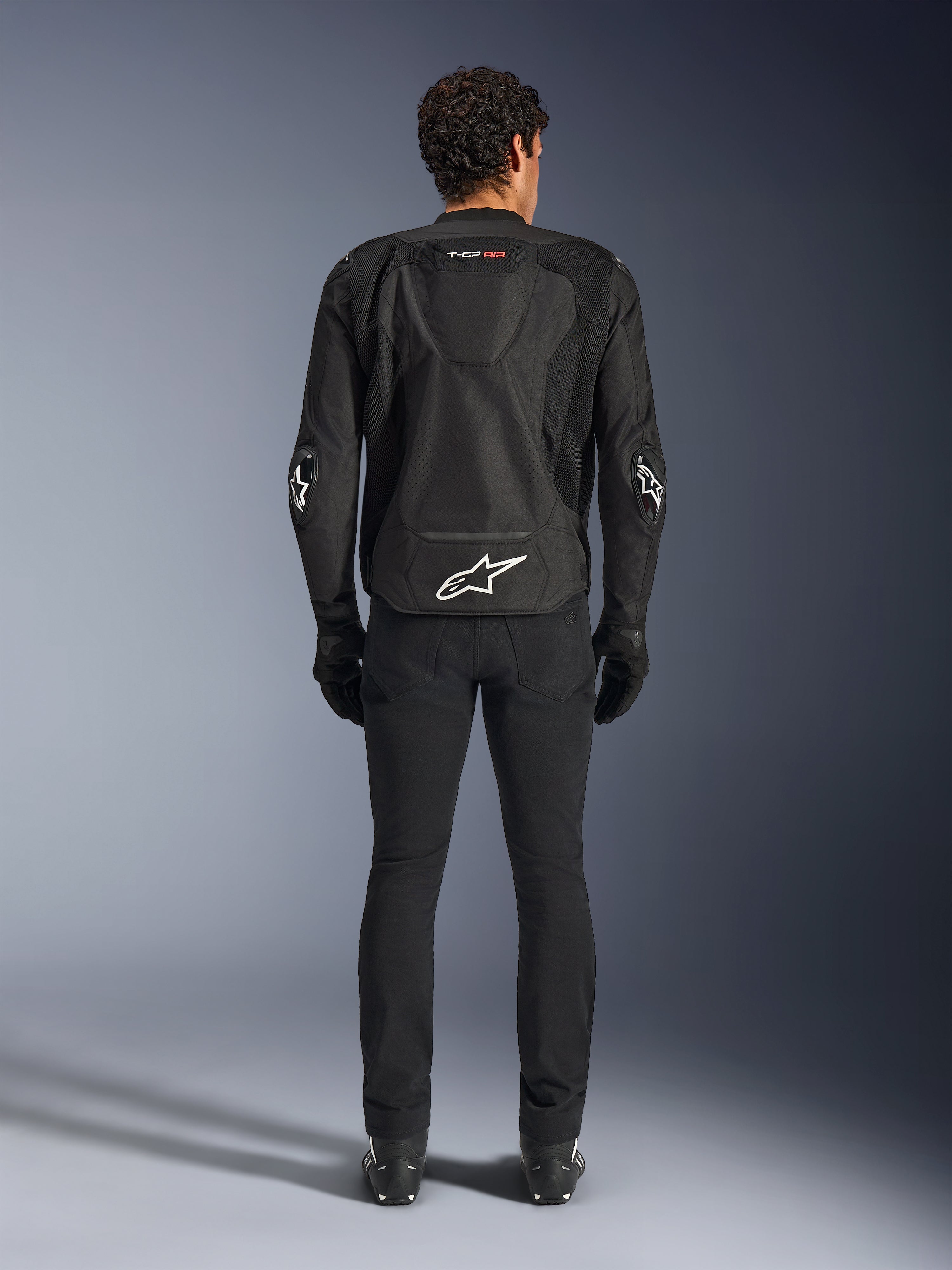 Casaco Alpinestars T-GP Air, Preto, vista traseira de um homem a usar o casaco de motociclista têxtil, com painéis de ventilação em malha, uma corcunda aerodinâmica e logótipos Alpinestars brancos na zona lombar e nos cotovelos, usado com calças de condução e luvas pretas