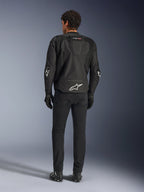 Casaco Alpinestars T-GP Air, preto, vista traseira no modelo, com painéis de ventilação em malha 3D e corcunda aerodinâmica, logótipo de estrela branco na zona lombar e cotovelos, usado com calças e luvas de motociclismo pretas, vestuário de condução desportiva.